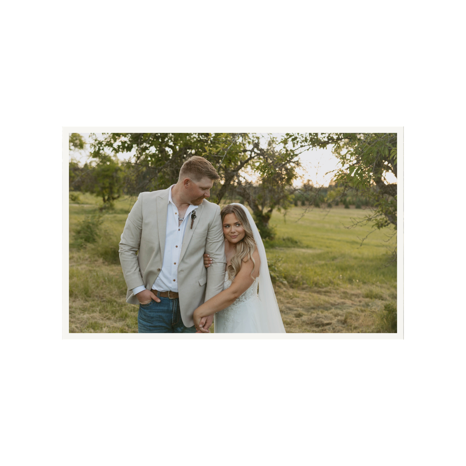 A couple in wedding attire stands in a lush green field, the groom in a tan suit and bride in a white gown.