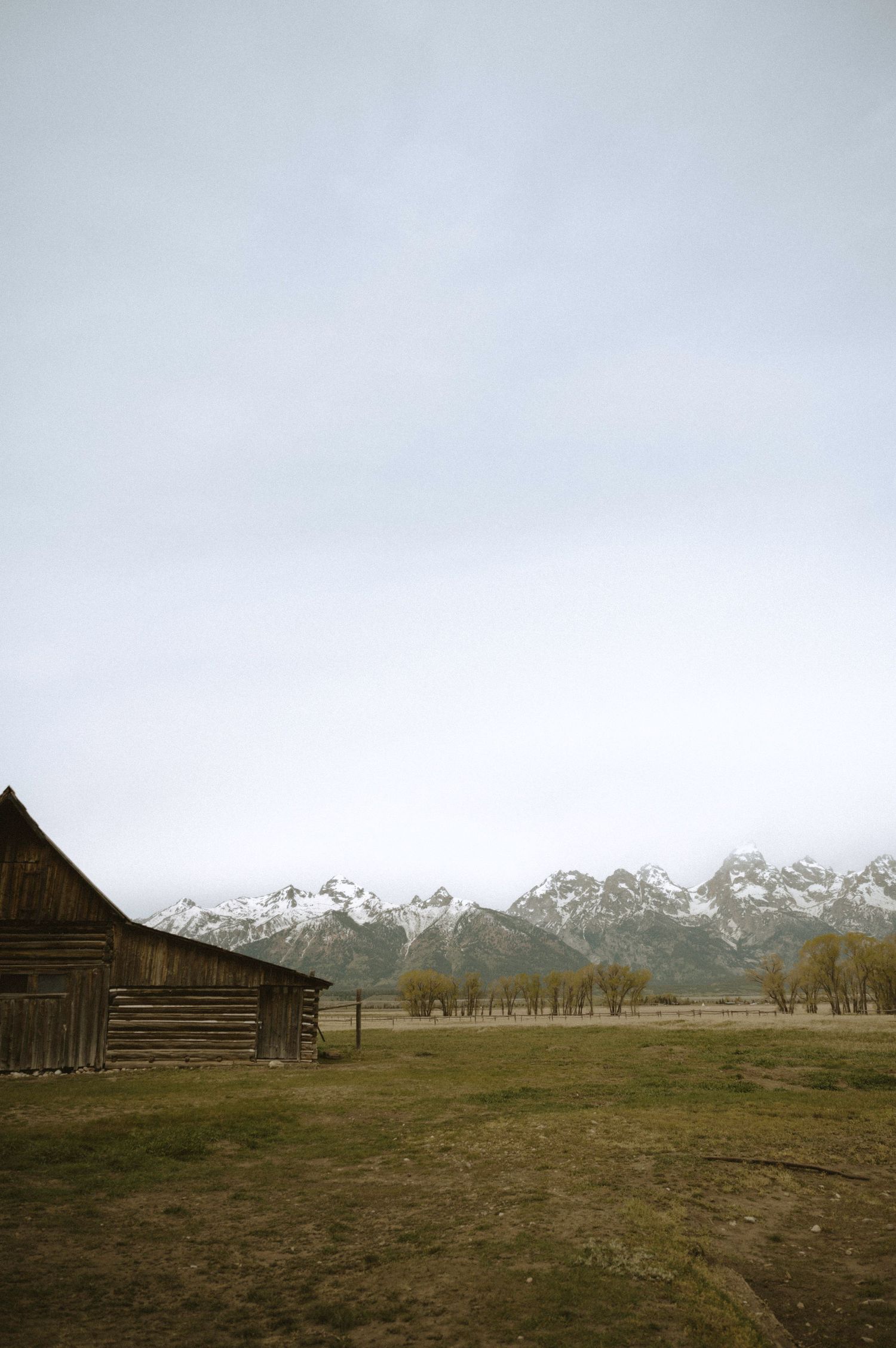 Rustic wooden barn in Grand Teton National Park with snow-capped mountains and overcast sky in background.