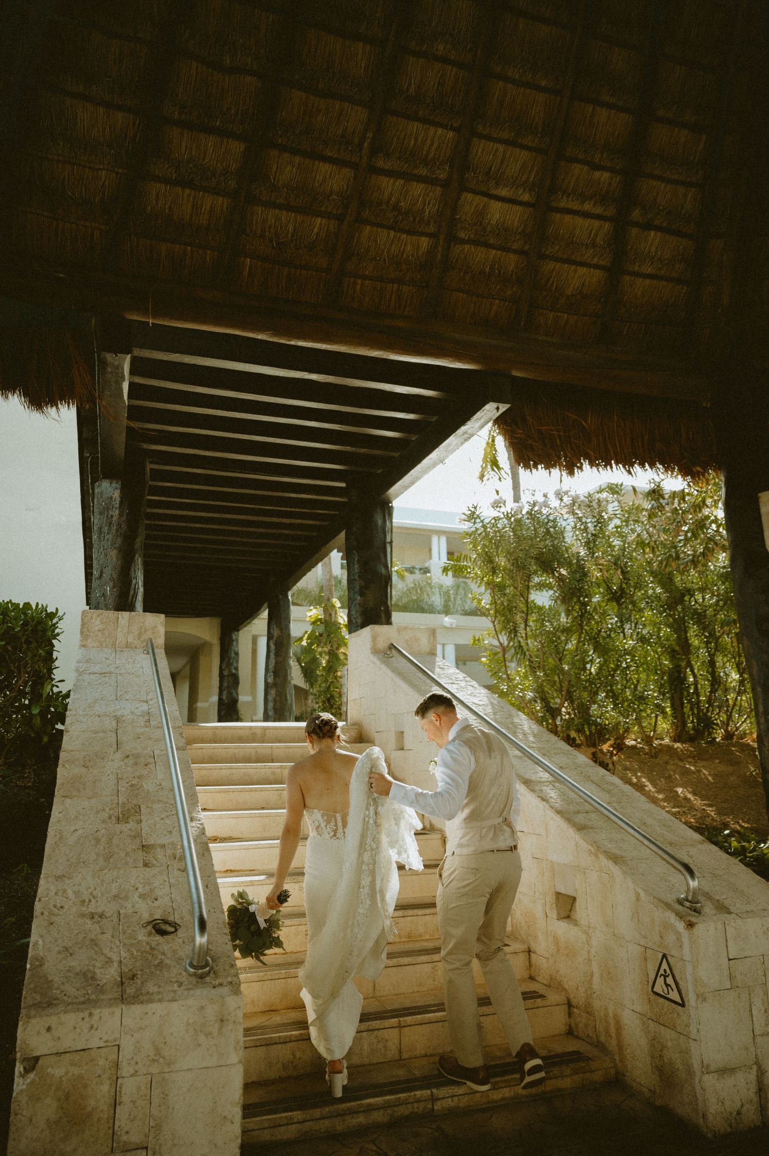 A couple in wedding attire ascends stone stairs under a thatched roof walkway at a tropical resort.