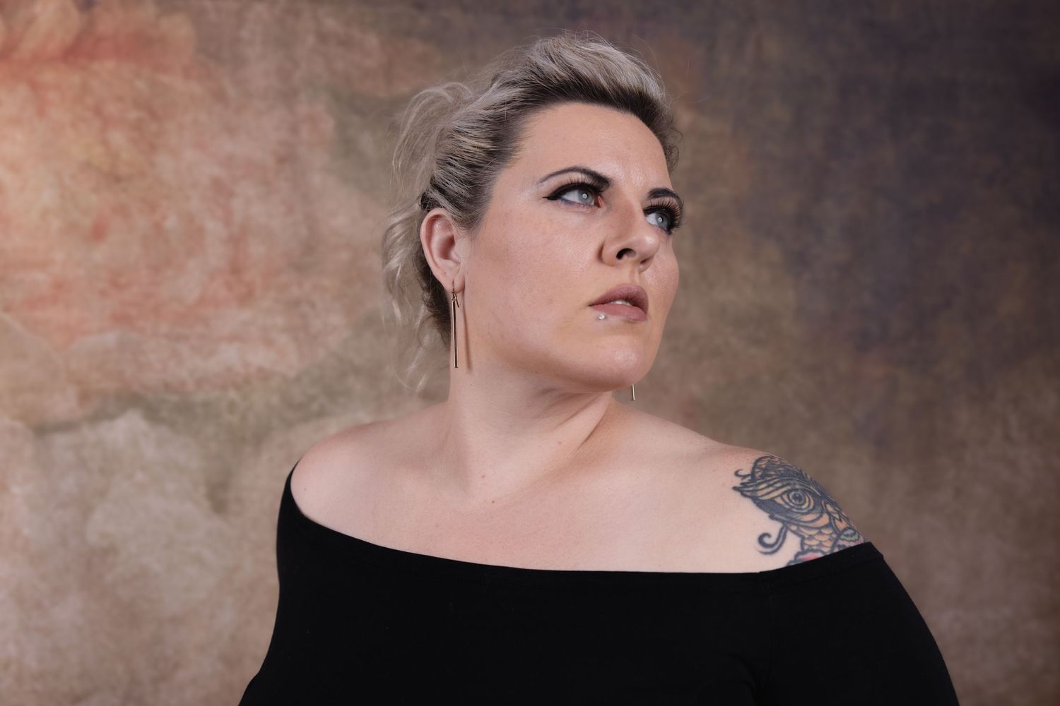 Dramatic portrait against textured wall showing shoulder tattoo and off-shoulder black top.