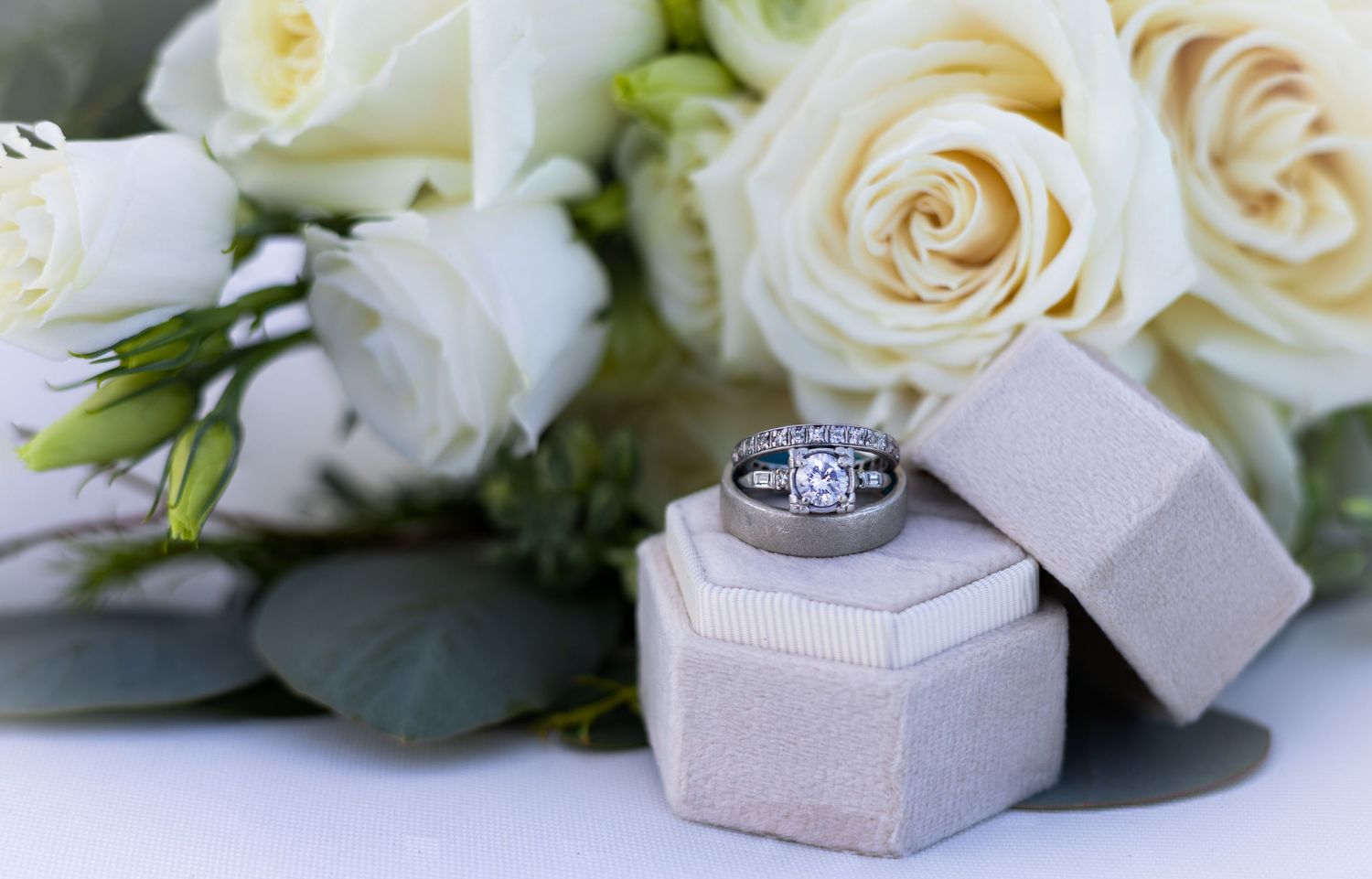 Wedding rings displayed in small hexagonal box with white roses and green foliage.