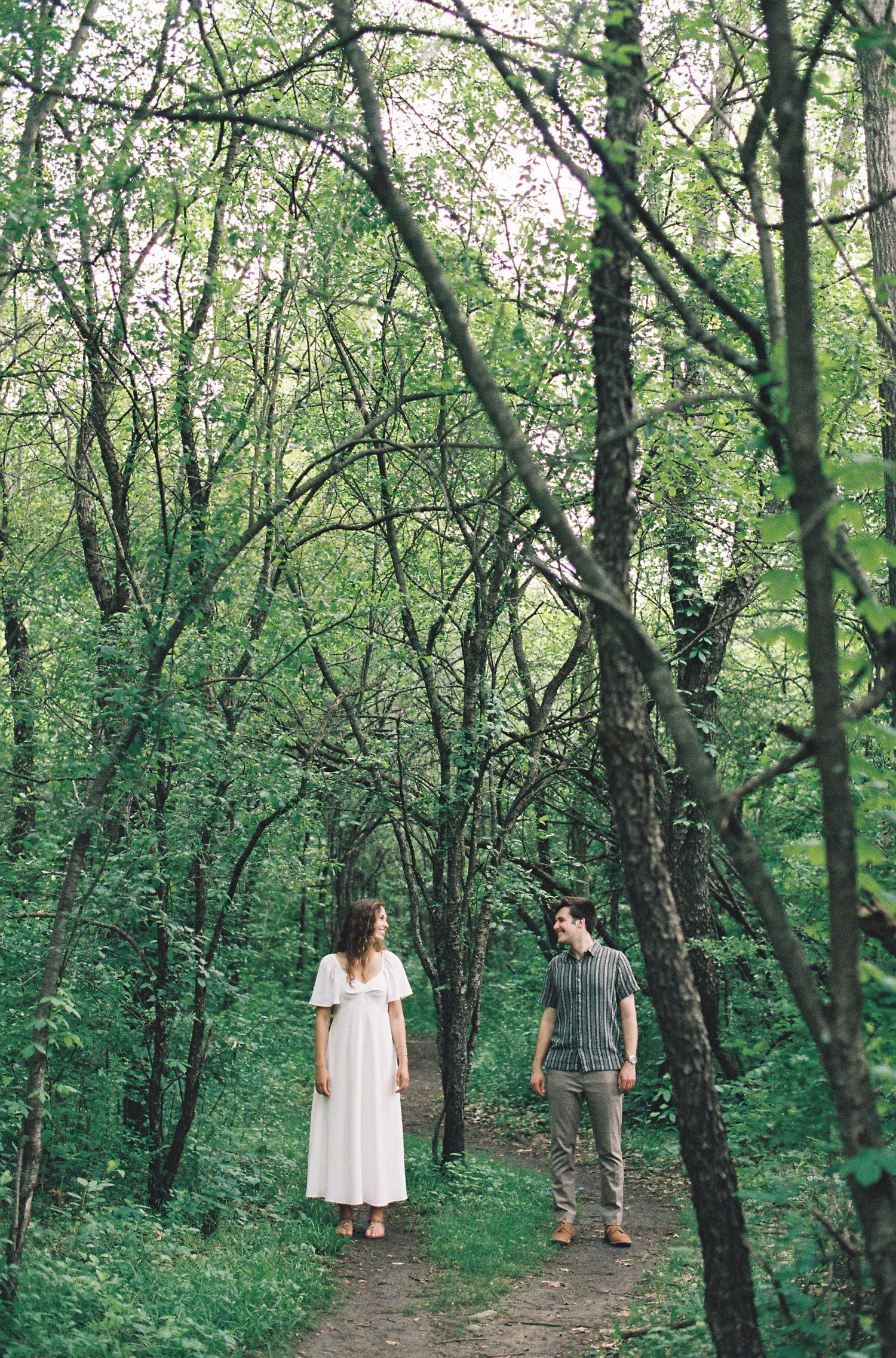 Couple standing on a forest path surrounded by lush green trees, woman in white dress, man in casual attire.