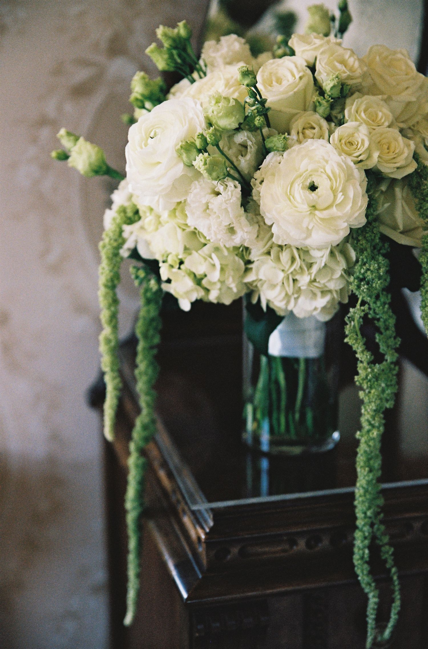 Elegant white floral bouquet with roses, hydrangeas, and green amaranthus in a glass vase on a dark wood table.