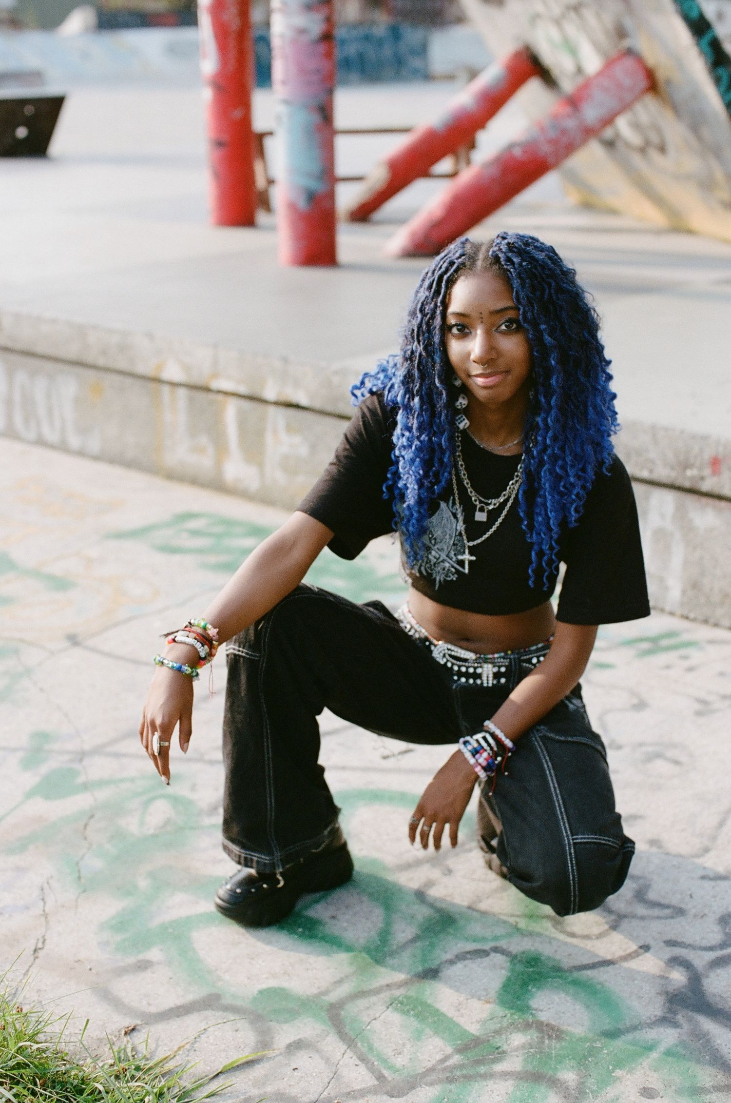 Young woman with blue braided hair in black crop top and wide-leg jeans crouching at an urban skate park.