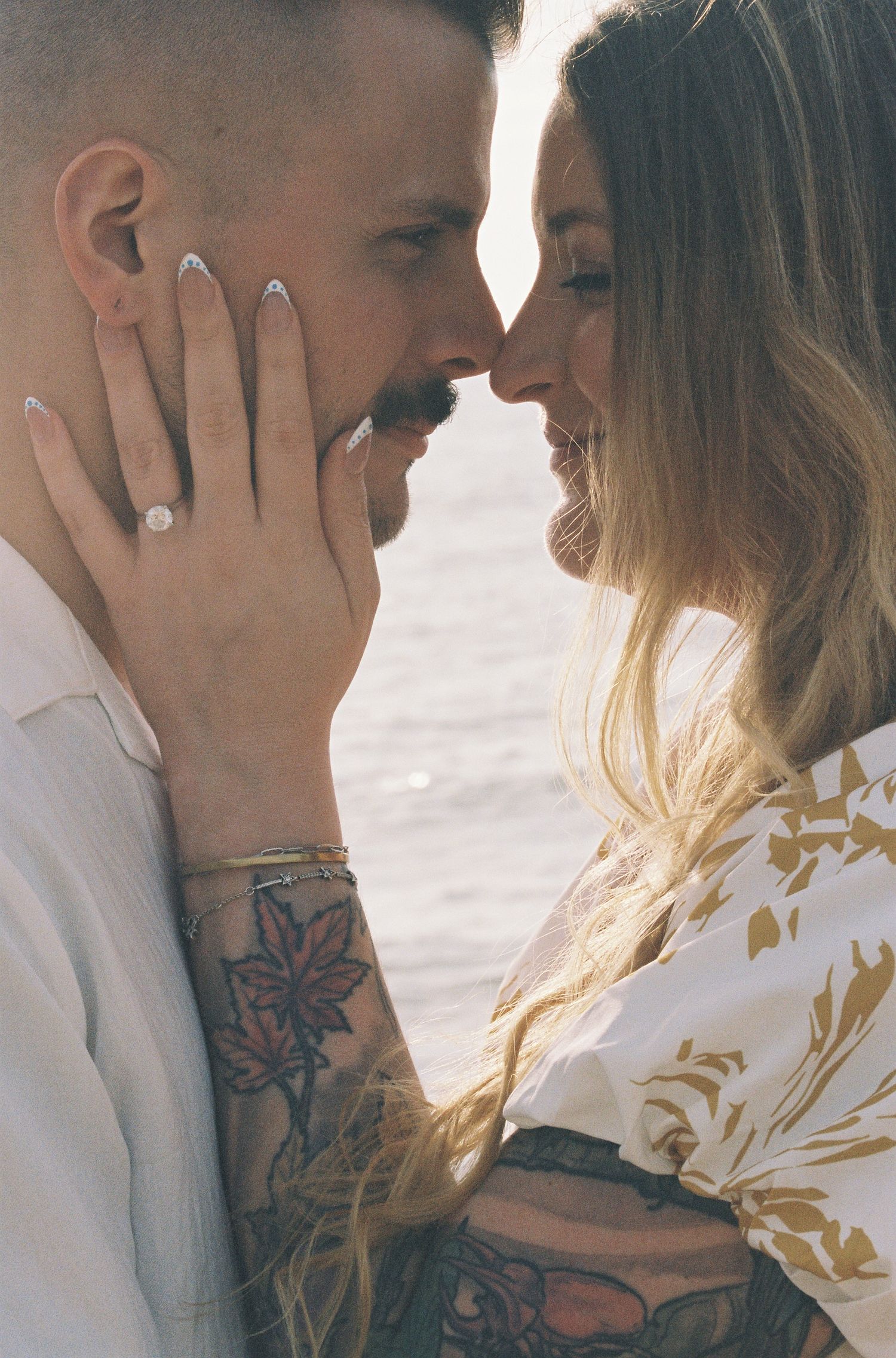 Couple sharing a romantic moment near water, woman showing engagement ring with tattoos visible on her arm.