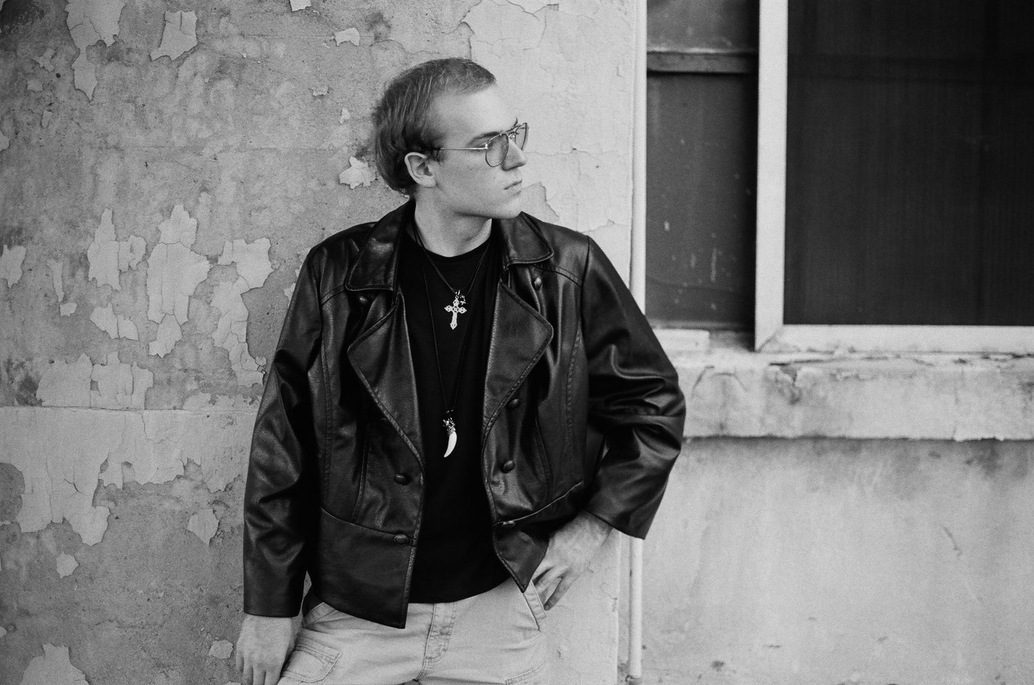 Man in leather jacket and glasses leaning against weathered wall, wearing cross necklace, black and white photo.