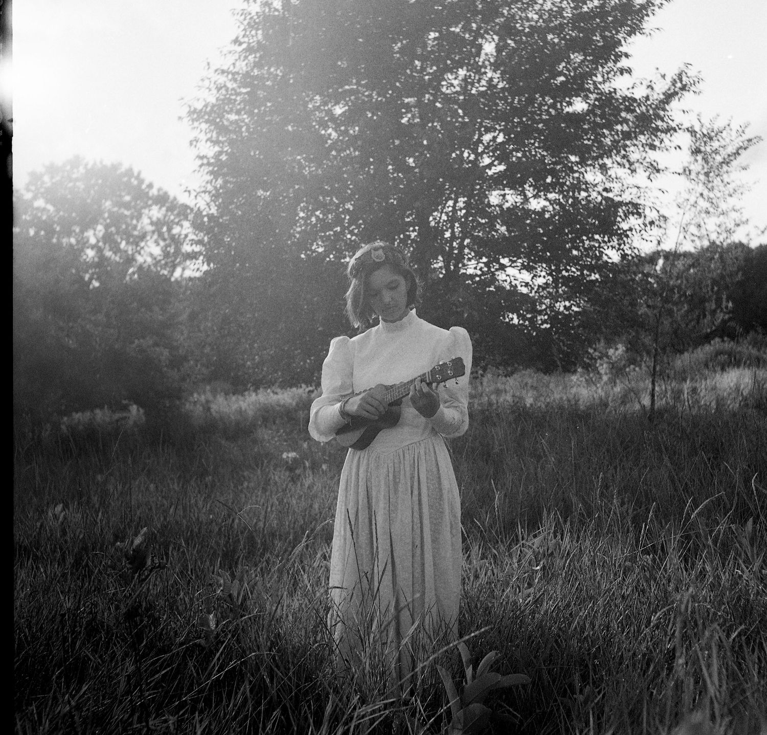 Vintage black and white photo of a young woman in a white dress playing ukulele outdoors in a field.