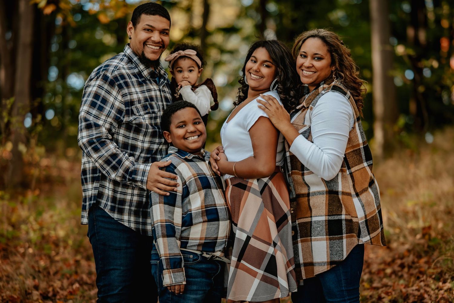 A family wearing coordinated plaid and neutral outfits poses together in an autumn forest setting during golden hour.