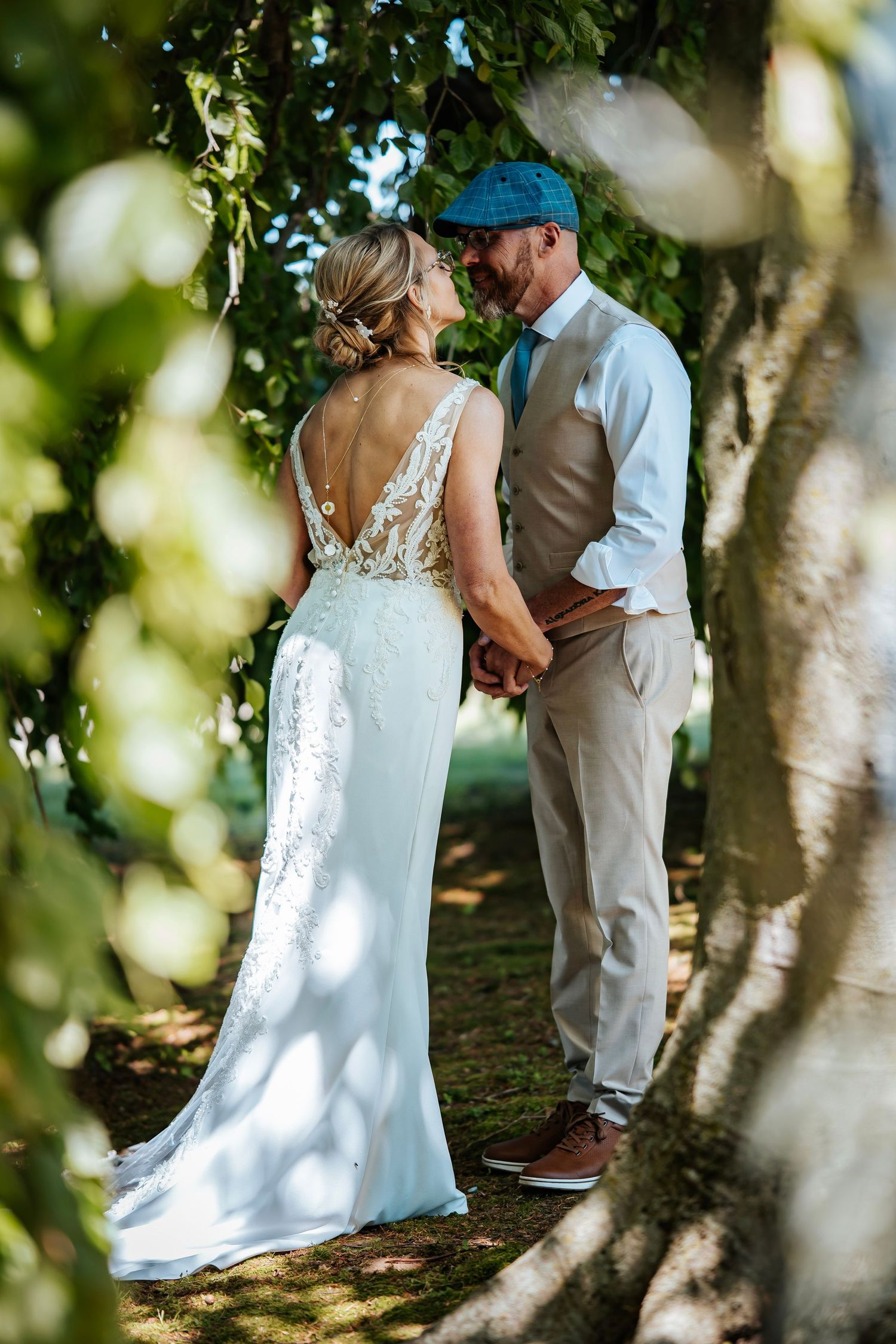 Bride and groom holding hands under trees, bride in backless lace gown, groom in tan vest and blue cap.