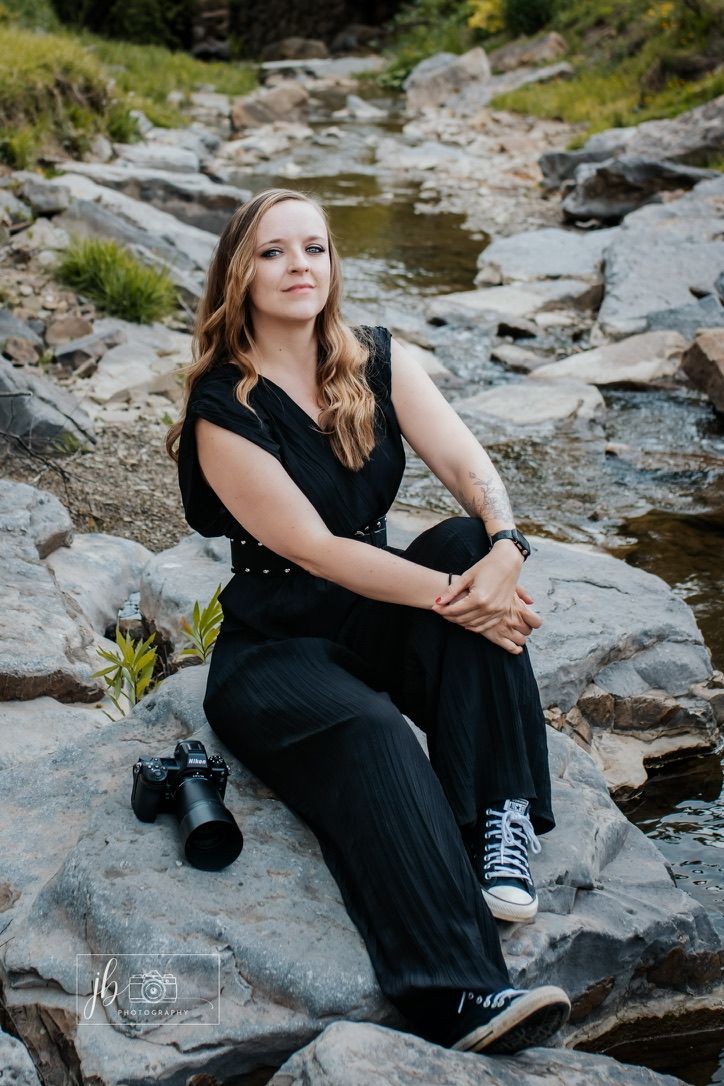 Woman in black jumpsuit sitting on rocks by a stream with a camera, posing for a professional outdoor portrait photo.