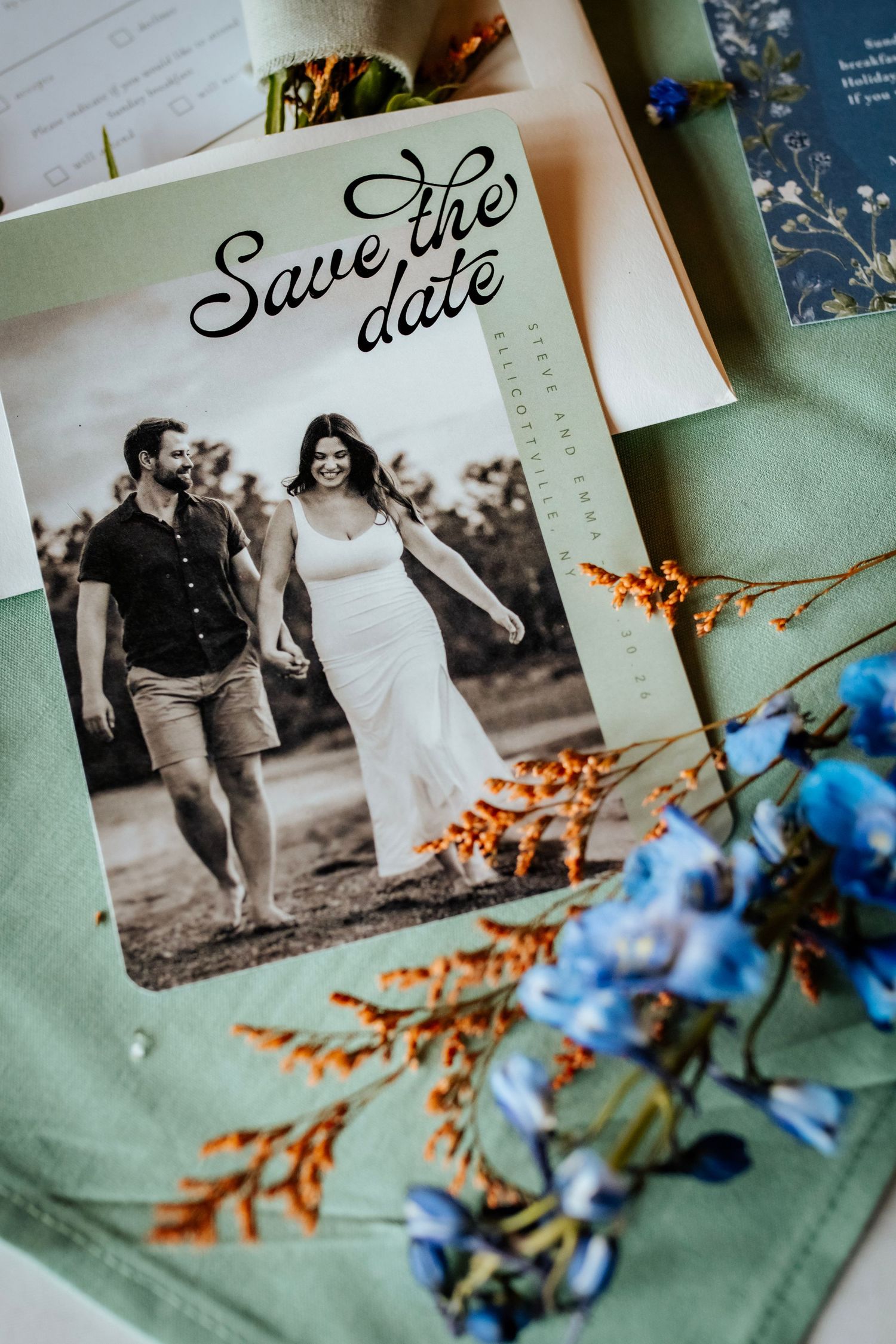 Save the date card with couple photo, blue flowers, and orange dried botanicals on sage green fabric.