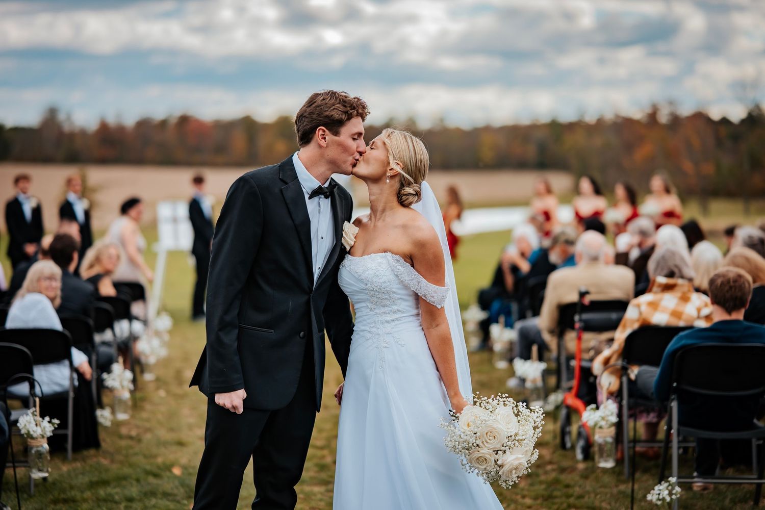 Bride and groom share a first kiss at outdoor fall wedding ceremony with guests seated in rows on a scenic field.