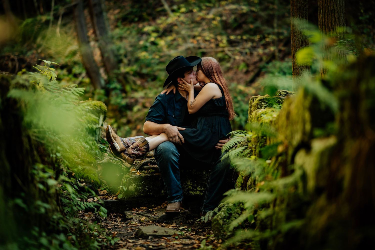Couple sharing a romantic kiss while sitting on mossy rocks in a lush green forest setting.