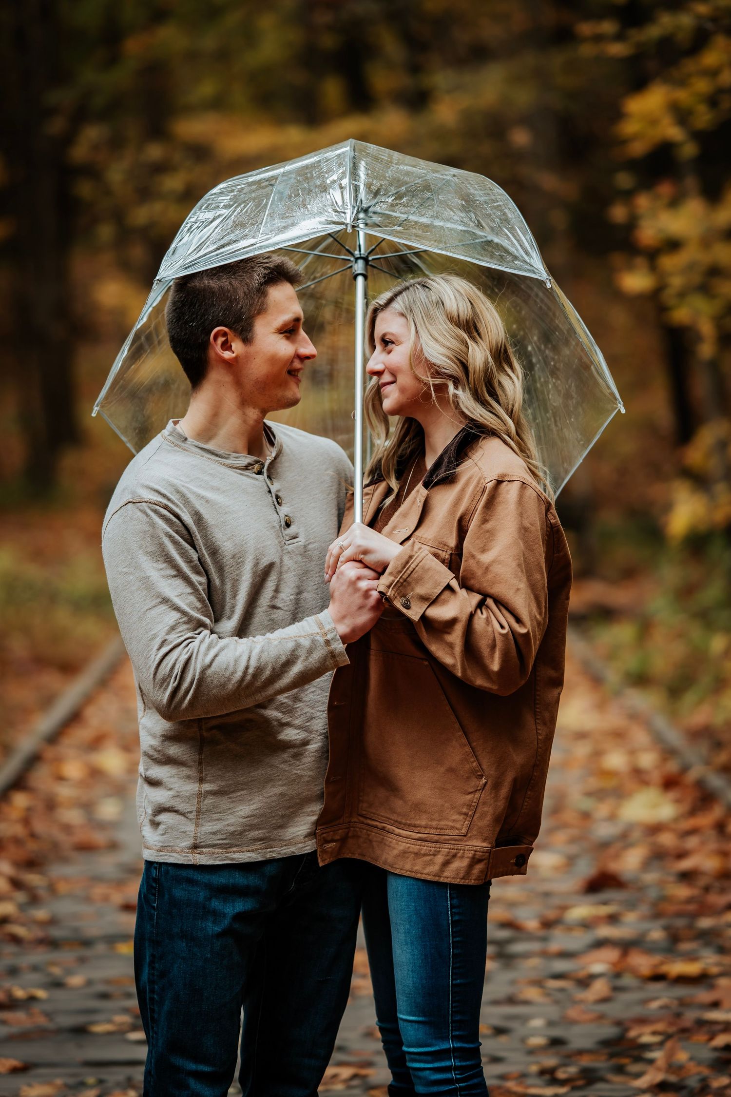 Couple sharing a romantic moment under a clear umbrella on autumn railway tracks surrounded by colorful fall foliage.