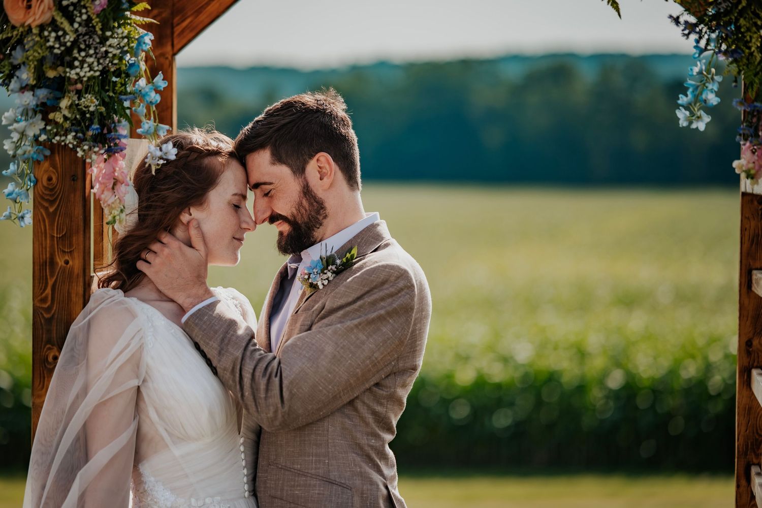 Bride and groom share a romantic moment under a floral arch with a green field backdrop at an outdoor wedding.