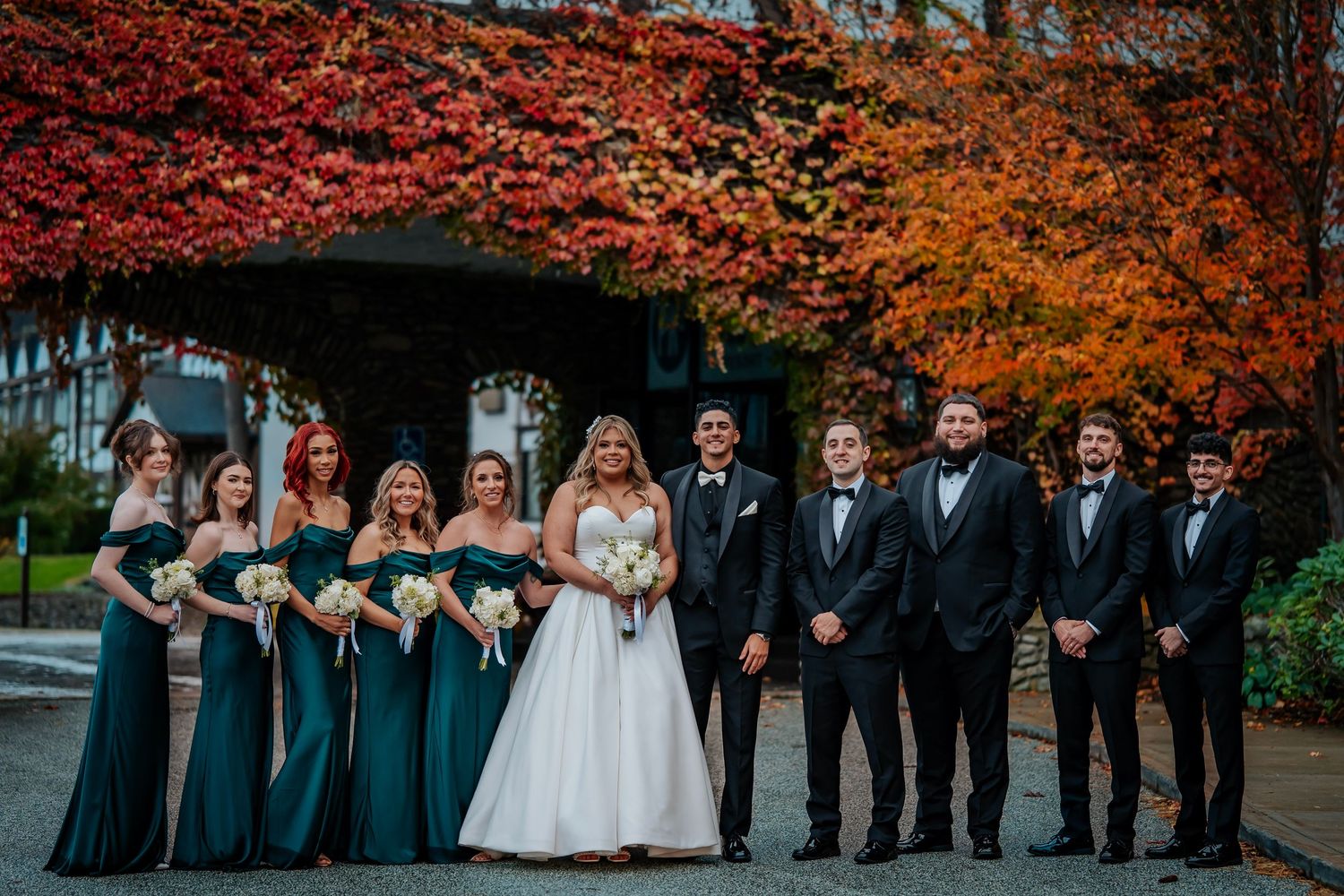 Wedding party stands together under vibrant autumn trees with red and orange foliage in the background.