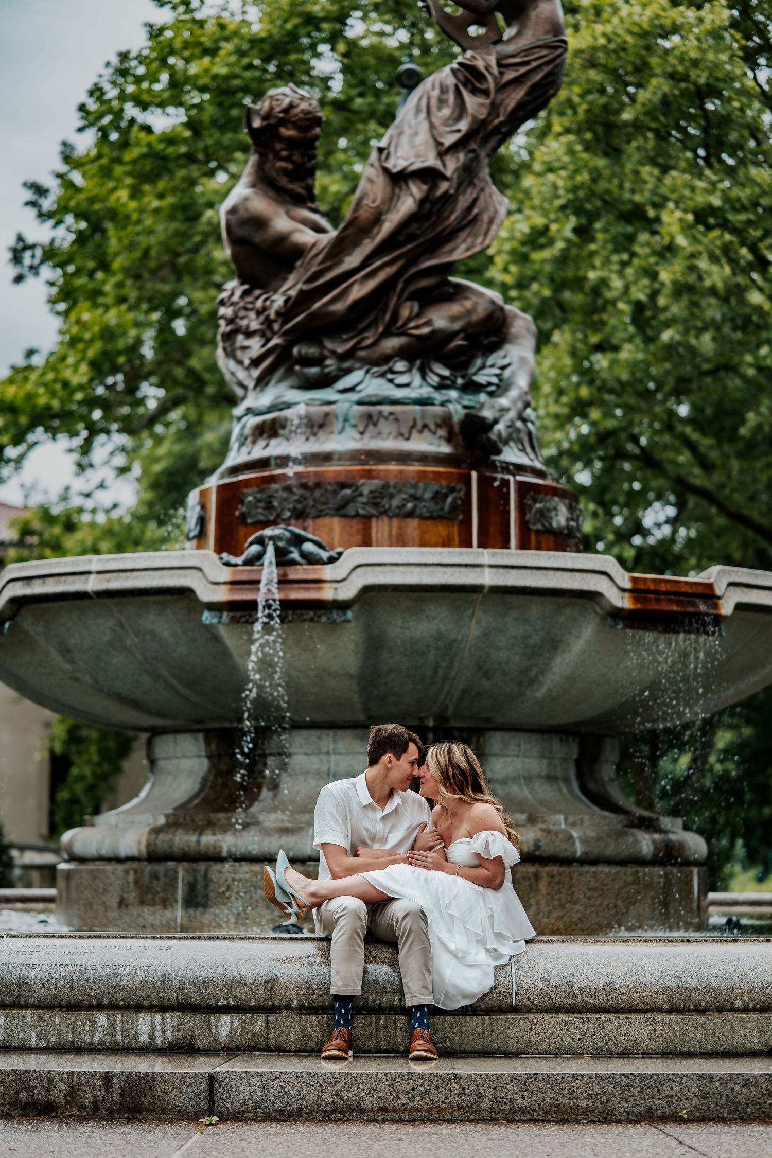 Couple kissing at ornate fountain with bronze sculpture in lush green park setting.