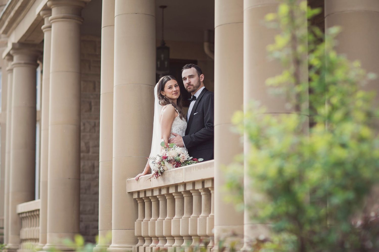 Bride and groom posing on elegant stone balcony with columns at a classic wedding venue.