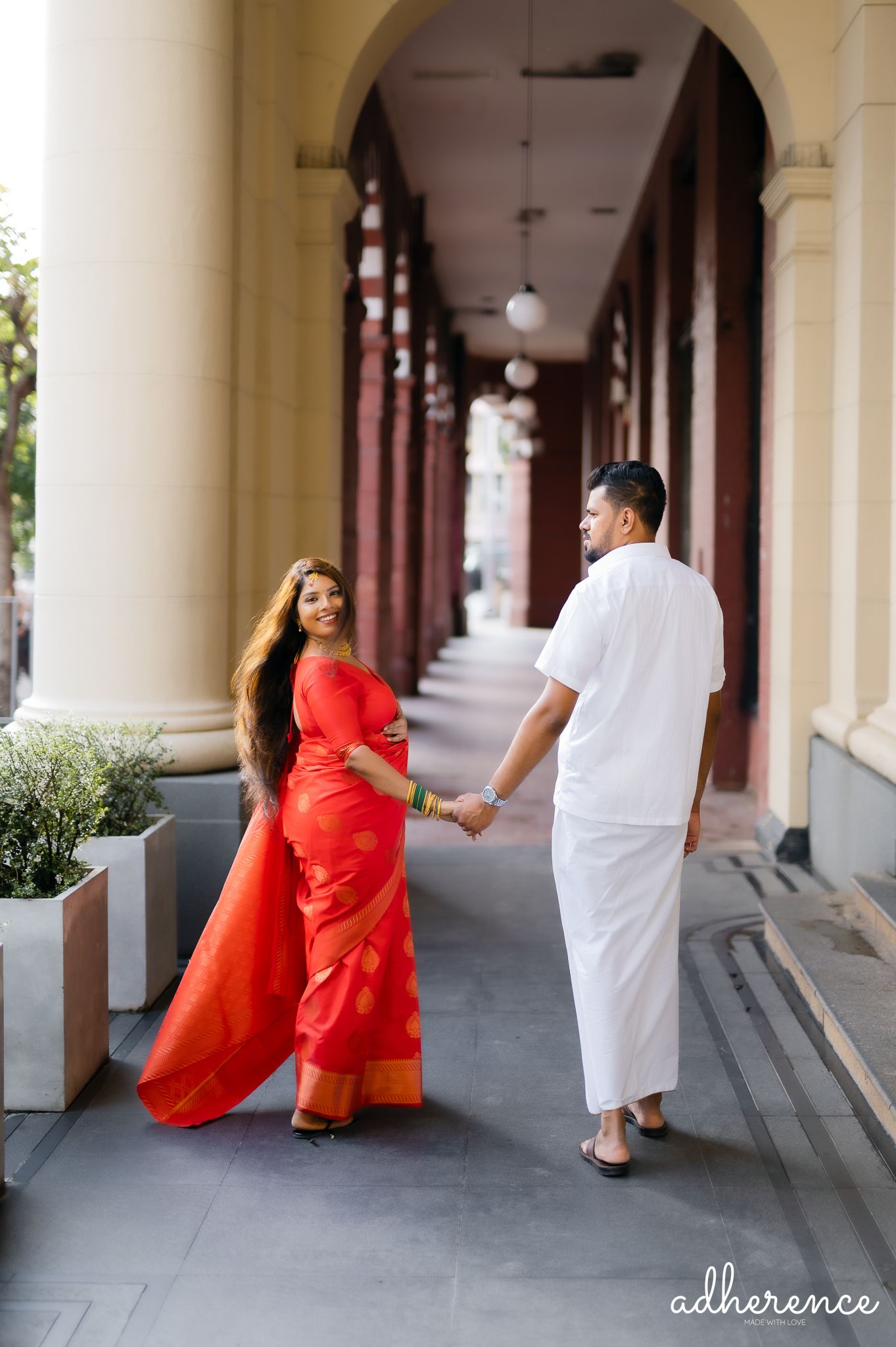 Couple walking in a colonnade with woman in flowing red saree and man in white.