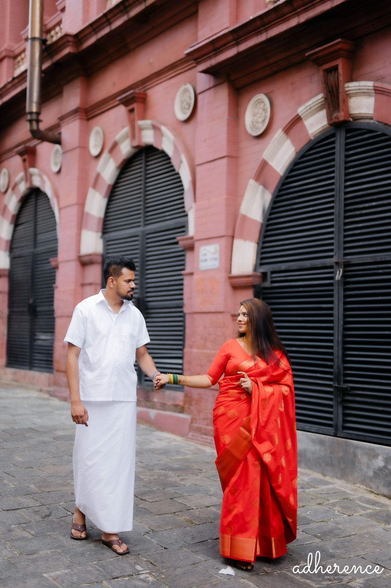 Couple in traditional white and red attire walking along pink colonial building with arched windows.