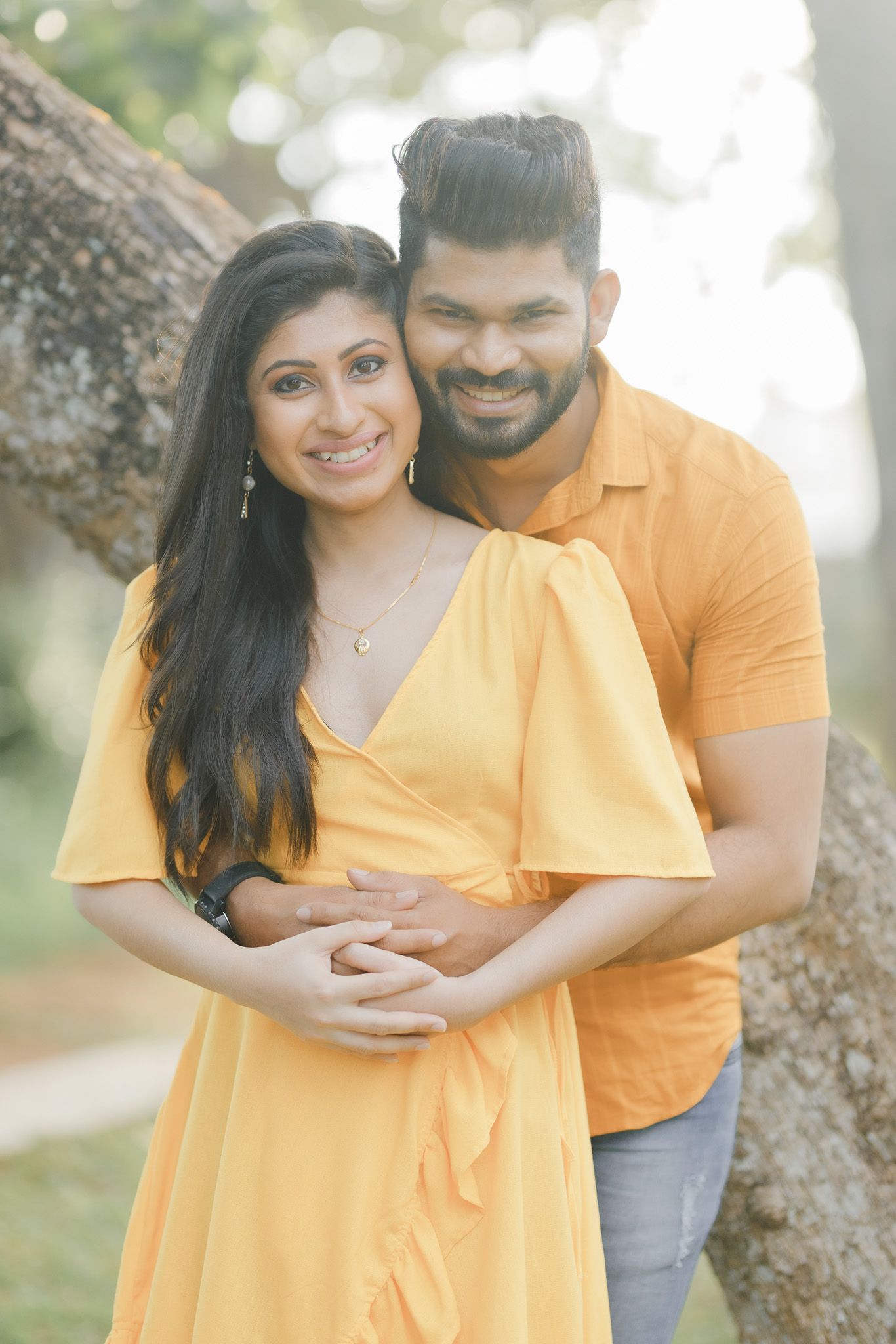 Couple in matching yellow outfits embrace by tree trunk in sunlit outdoor setting.