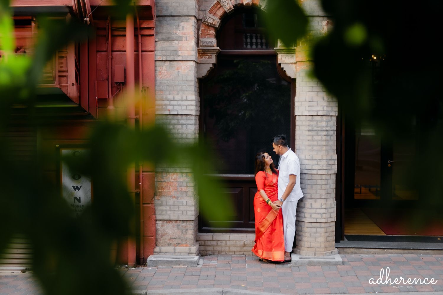 Couple in traditional attire poses beneath historic archway of pink building.