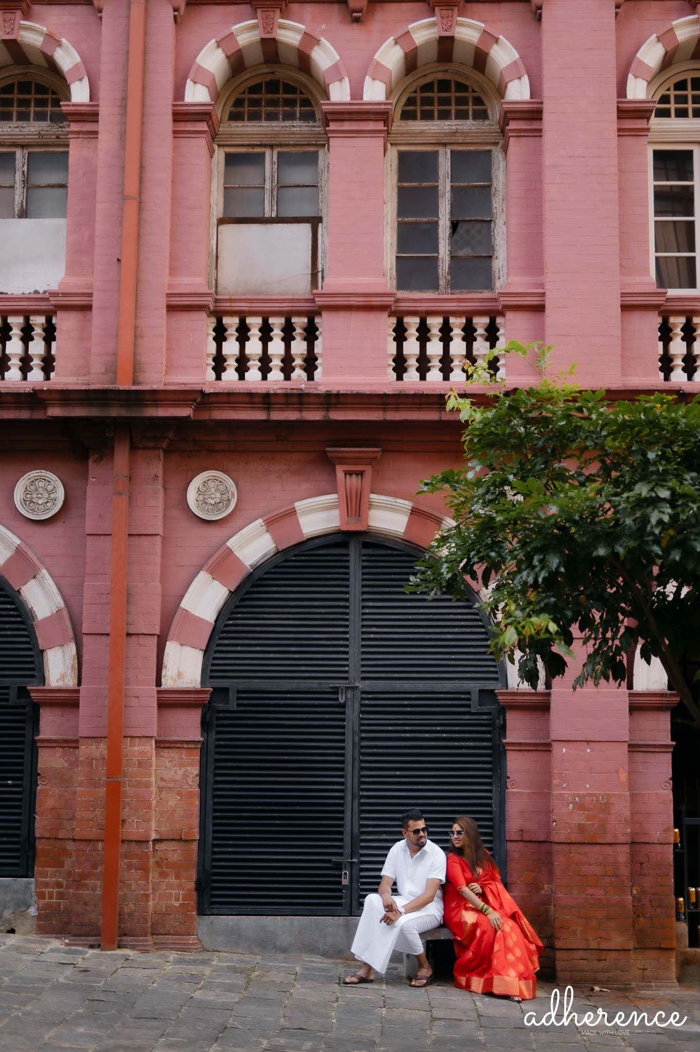 Couple in traditional attire sitting by pink colonial building with ornate architecture.