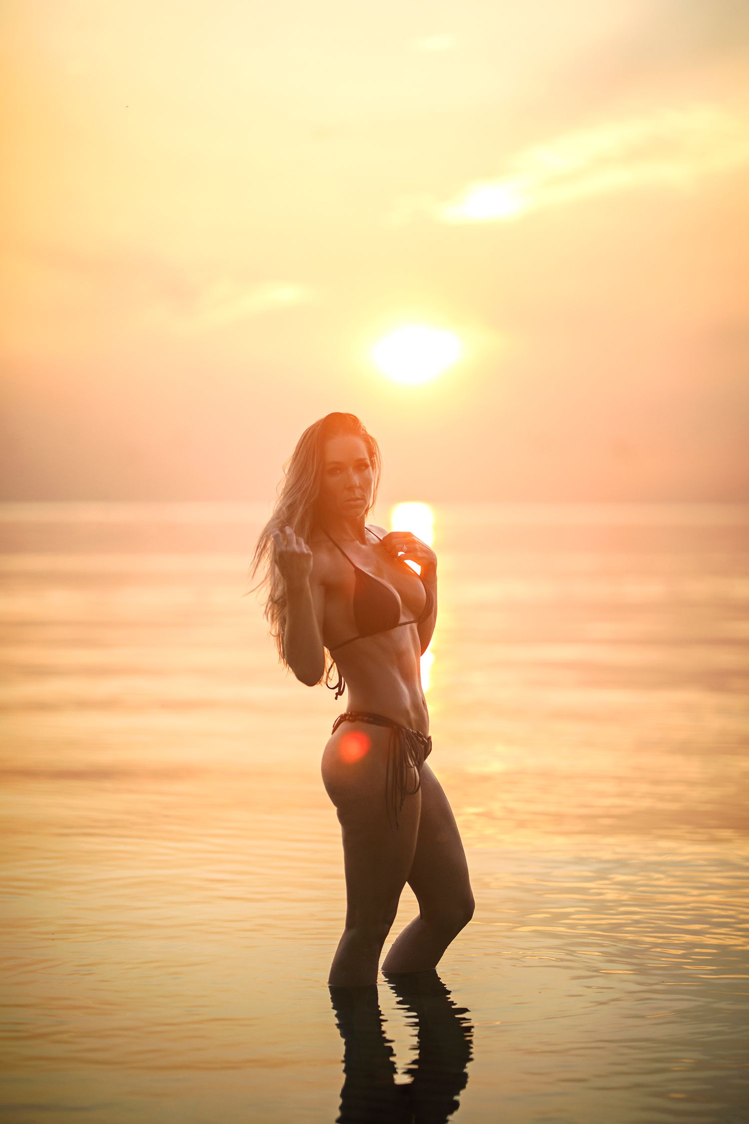 A woman in a bikini stands in shallow ocean water during a stunning golden sunset, with warm light reflecting off the sea.