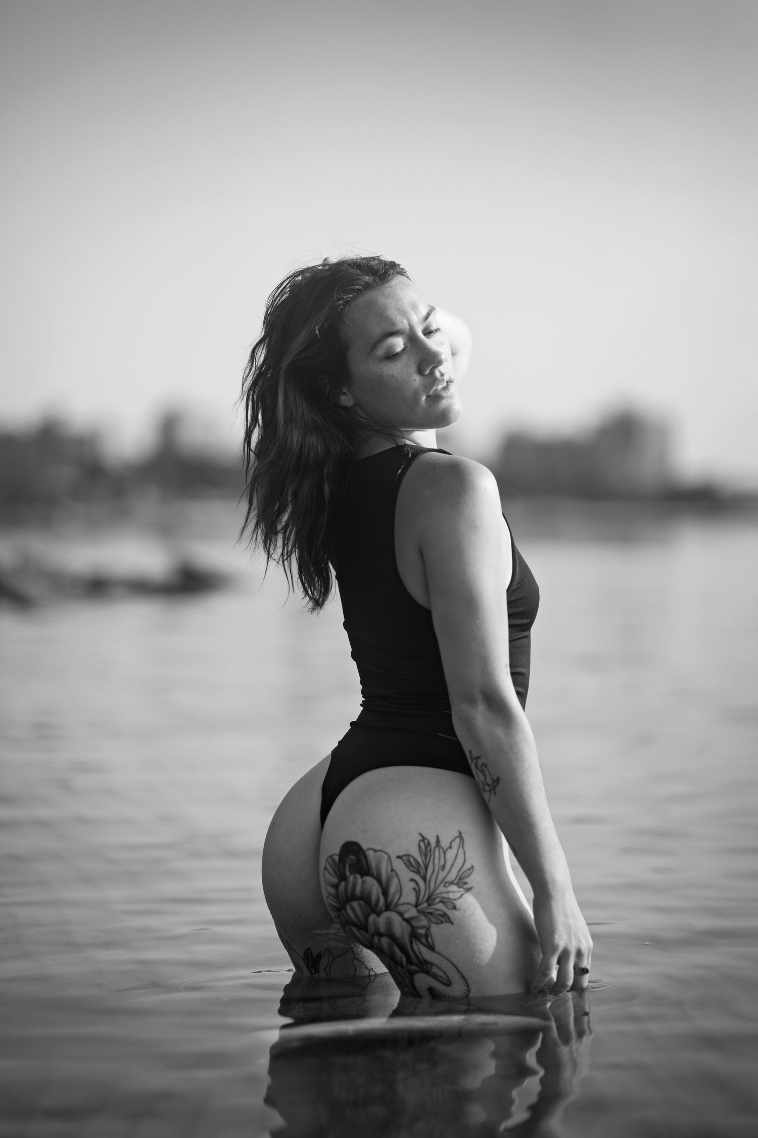 Black and white photo of a tattooed woman in a swimsuit standing in water, posing with eyes closed.