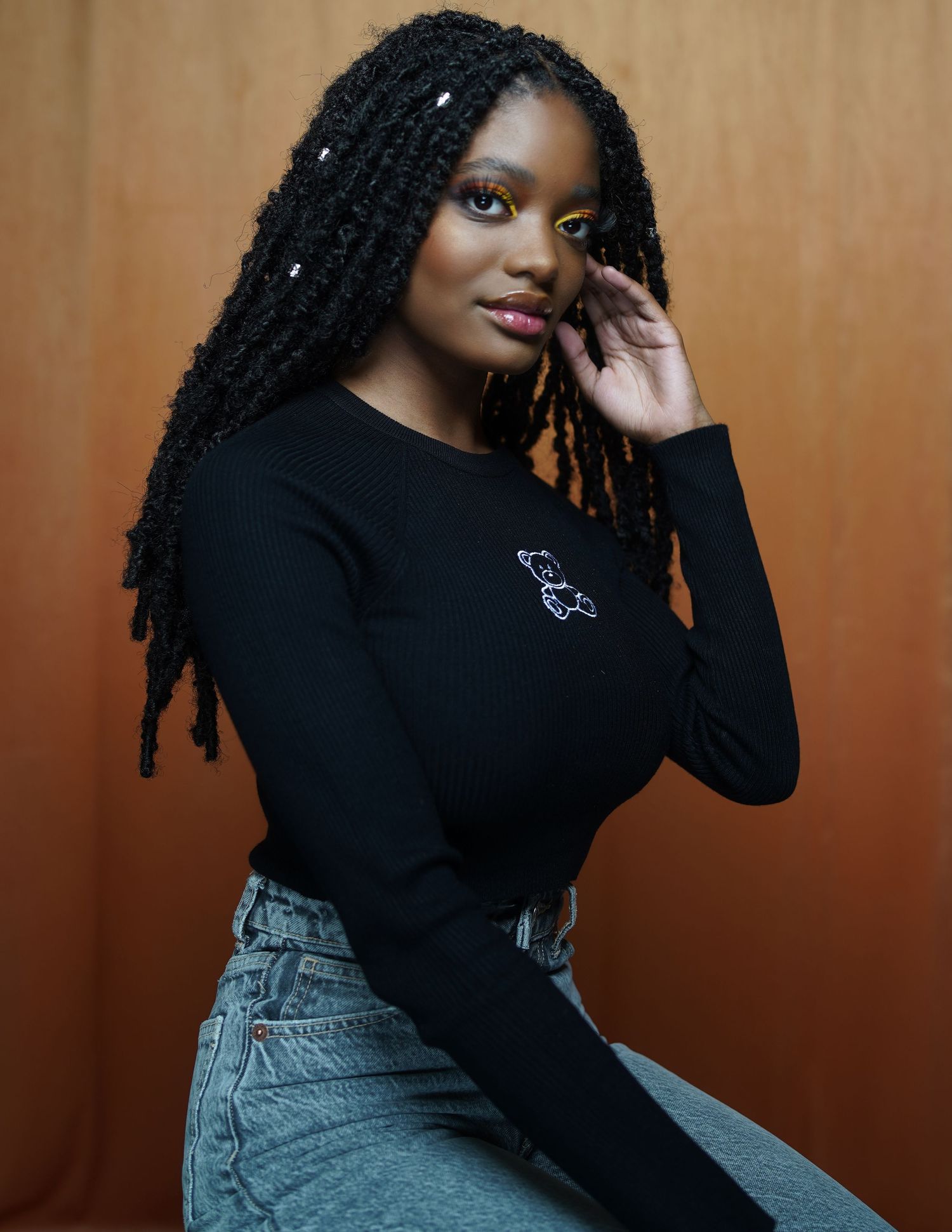 Young Black woman with braided locs wearing black long-sleeve top and jeans posing against warm background.