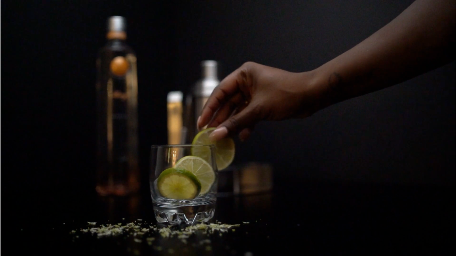 Hand placing lime slice into glass with premium spirit bottles in dark moody background.
