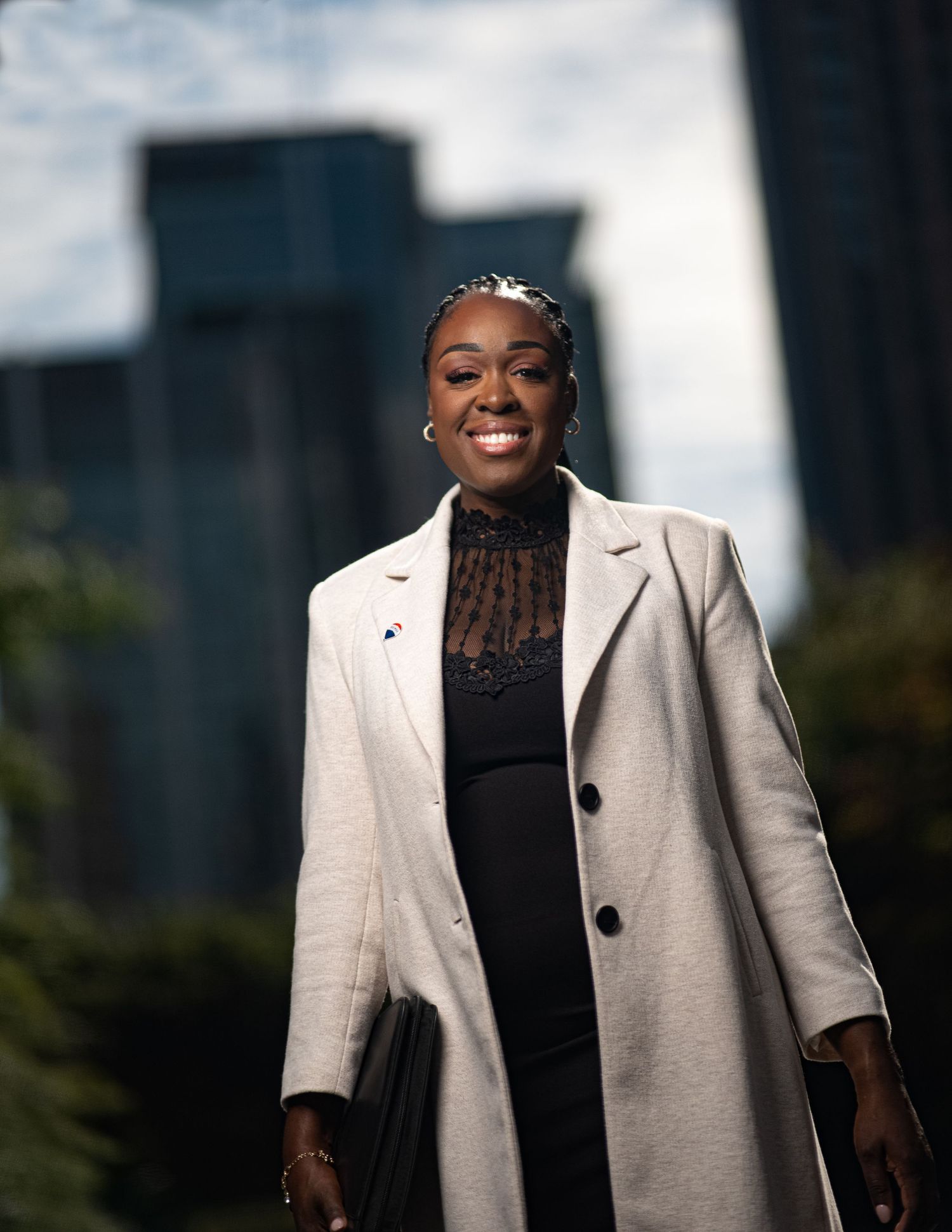 Smiling Black woman in white coat walking outdoors with city skyscrapers in background.