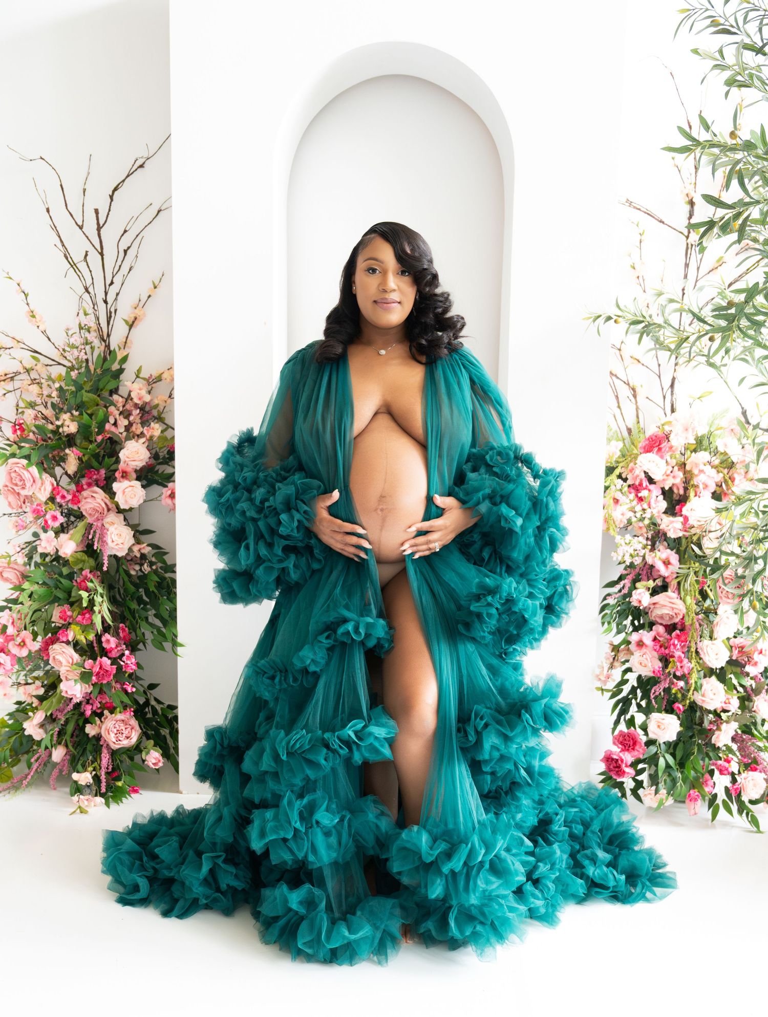 An elegant emerald green tulle robe with dramatic ruffles is photographed in a white archway surrounded by pink floral arrangements.