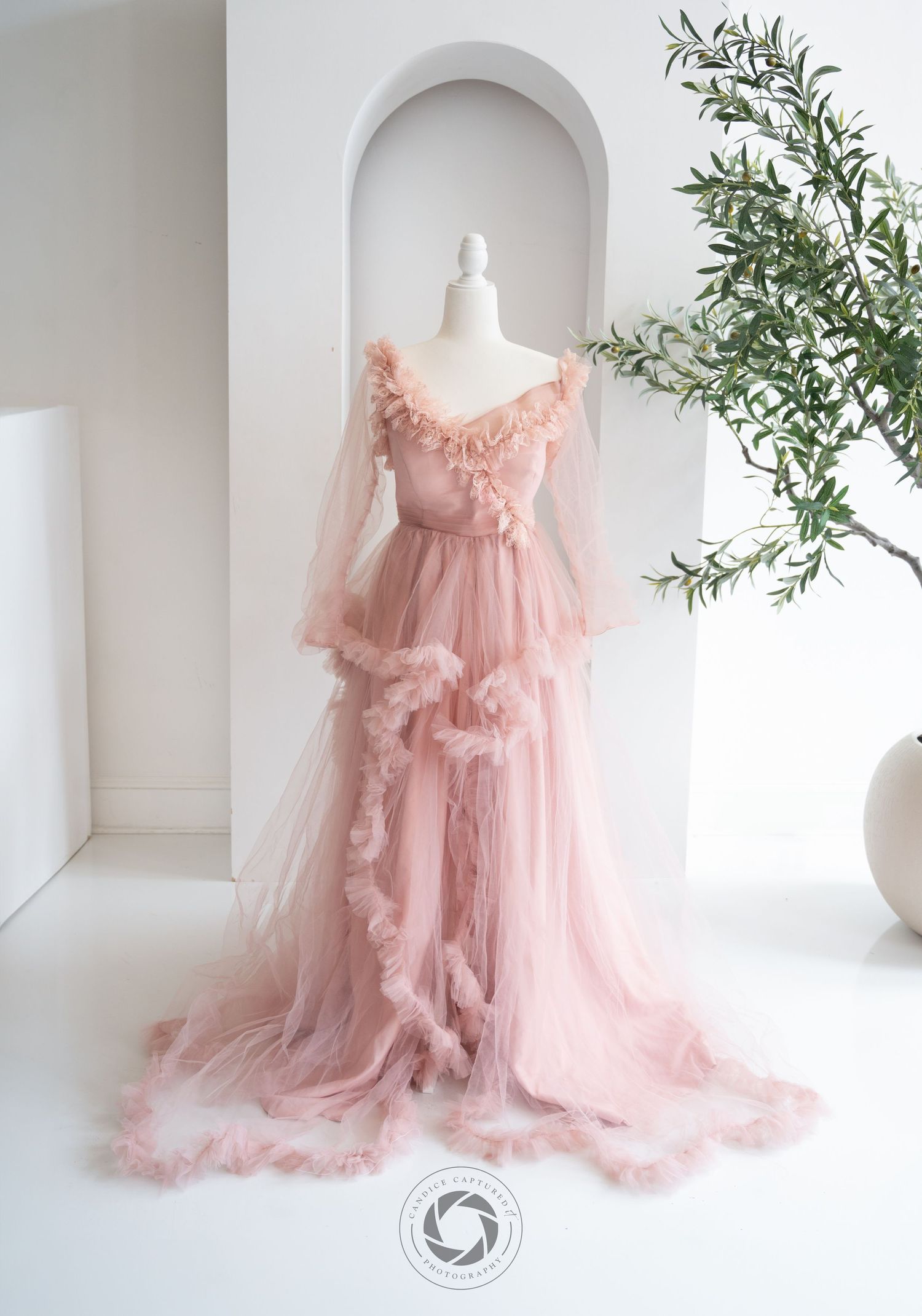Ethereal blush pink tulle gown with ruffled details and long bell sleeves displayed on mannequin against white arch wall.