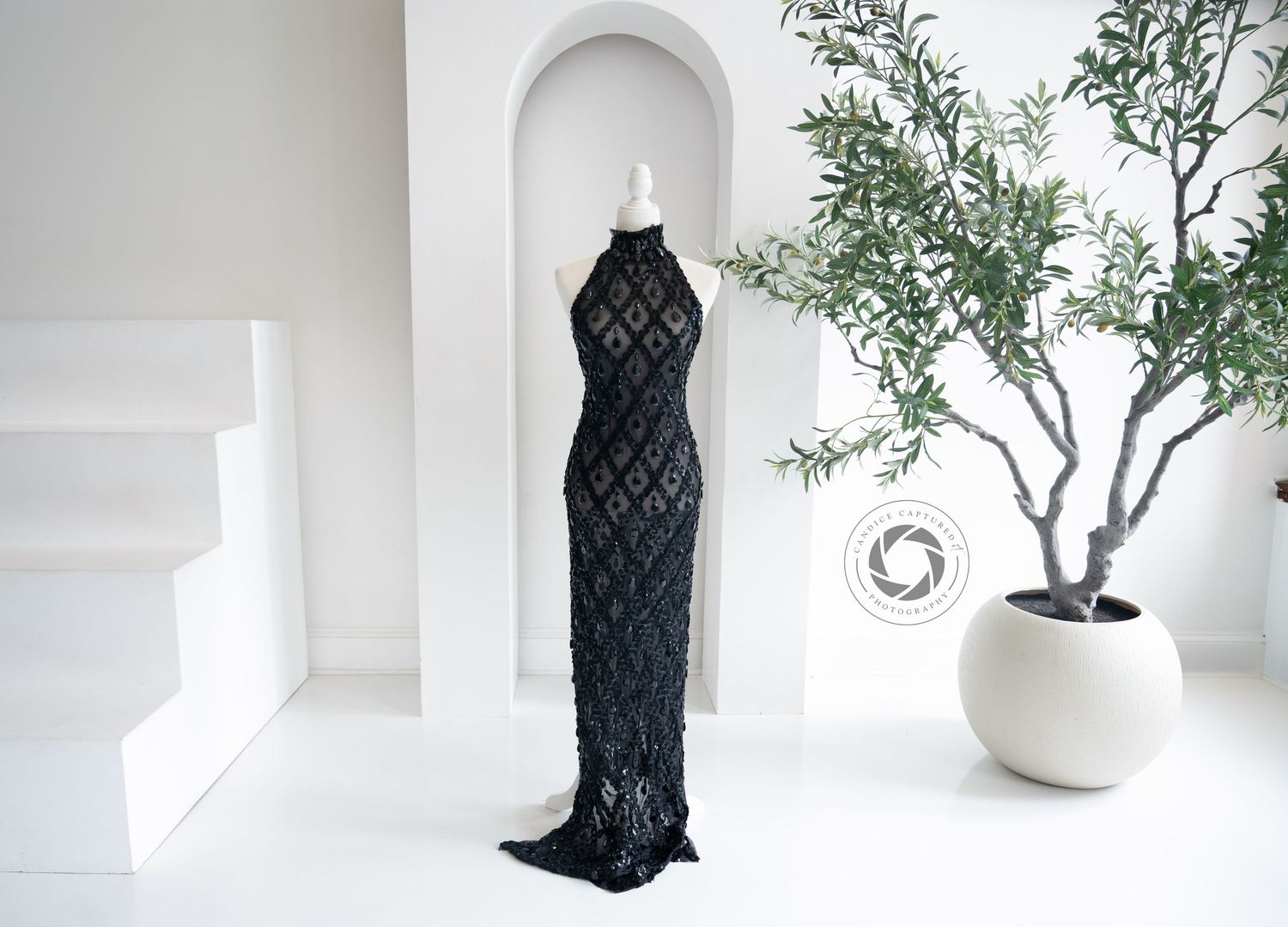 Elegant black sequined evening gown displayed in white minimalist space with arched niche and potted olive tree.