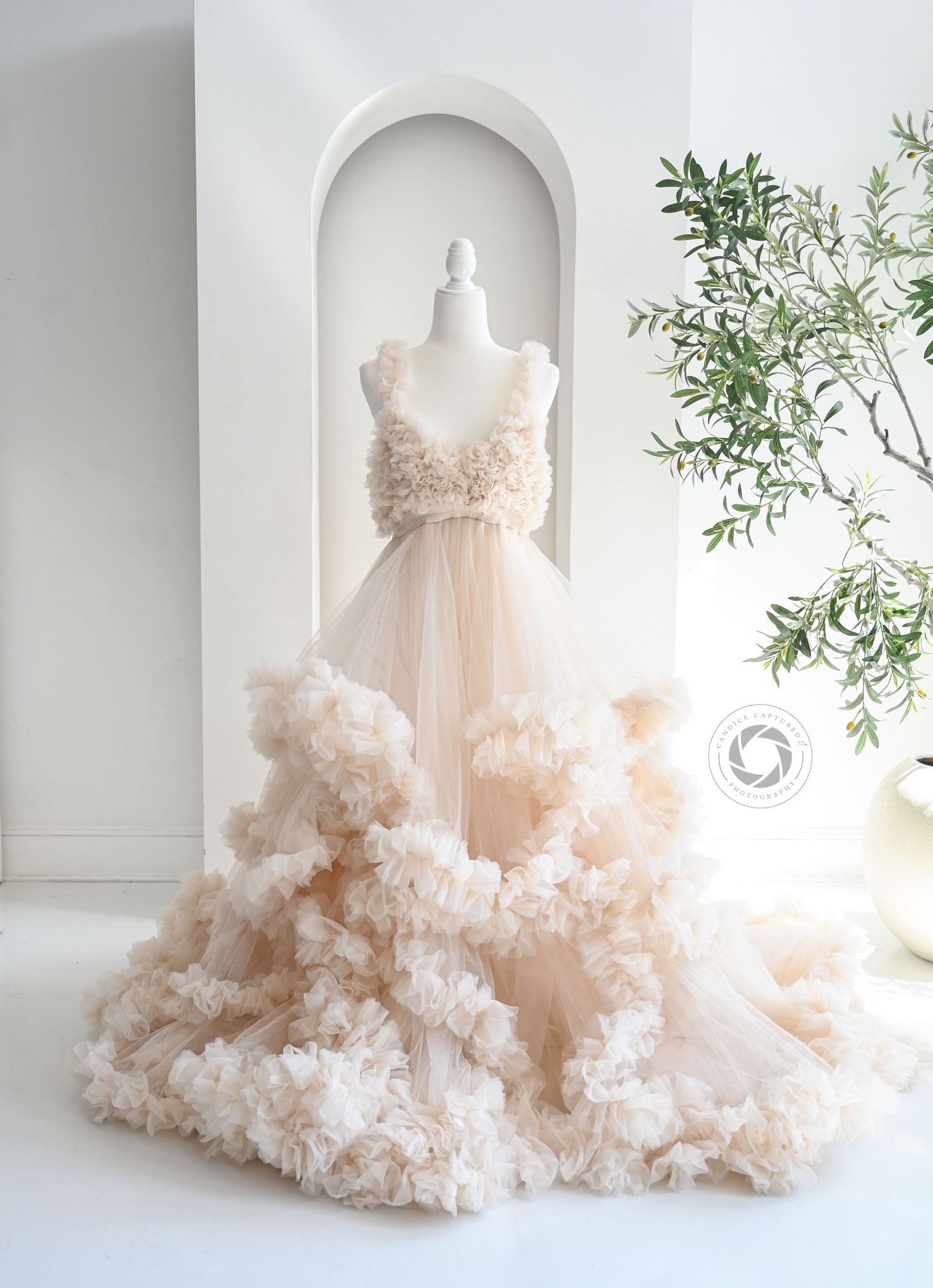 A luxurious blush pink tulle dress with ruffled layers and cloud-like texture displayed in a bright white studio setting.
