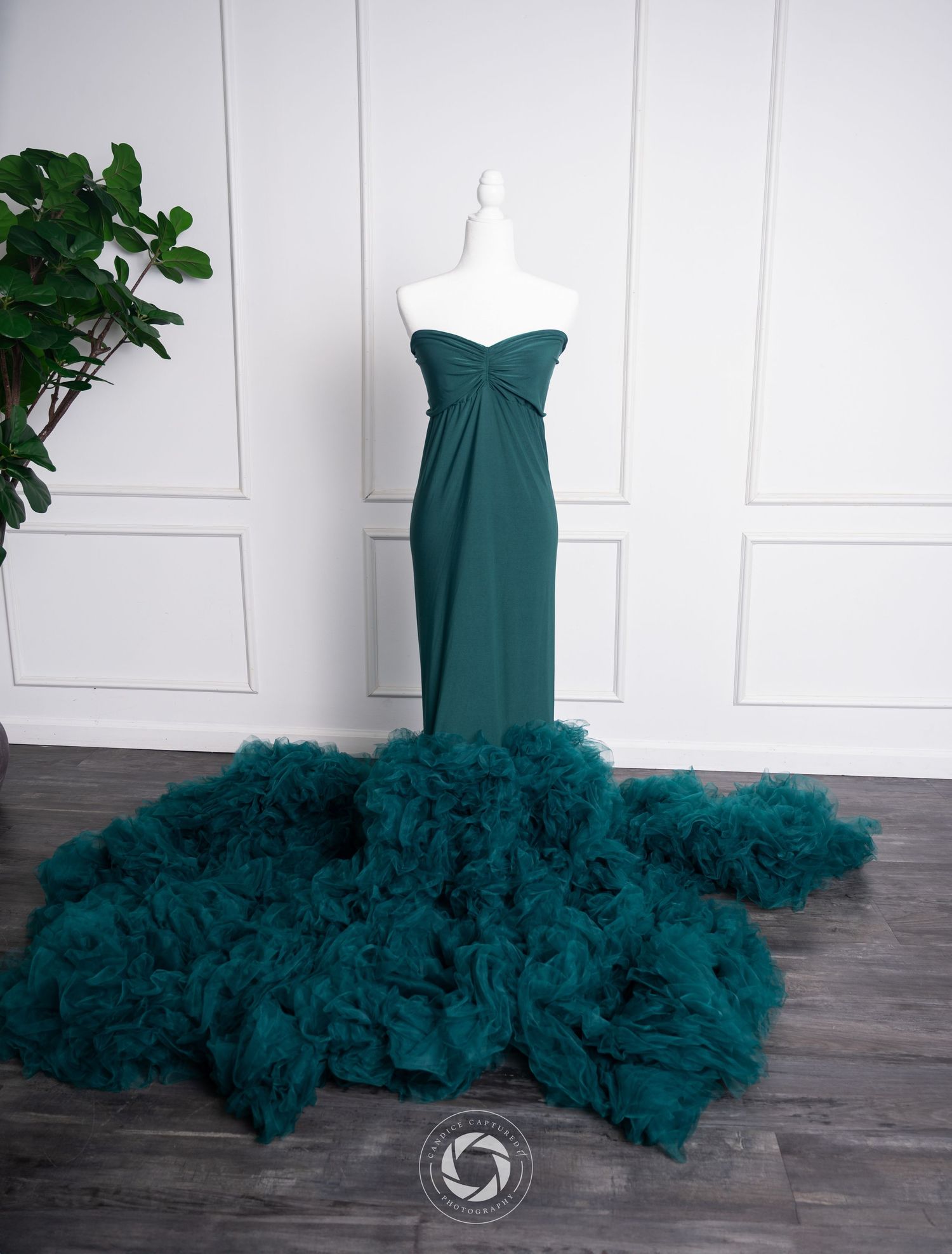 Elegant teal mermaid gown with dramatic tulle train displayed against white paneled wall next to green potted plant.