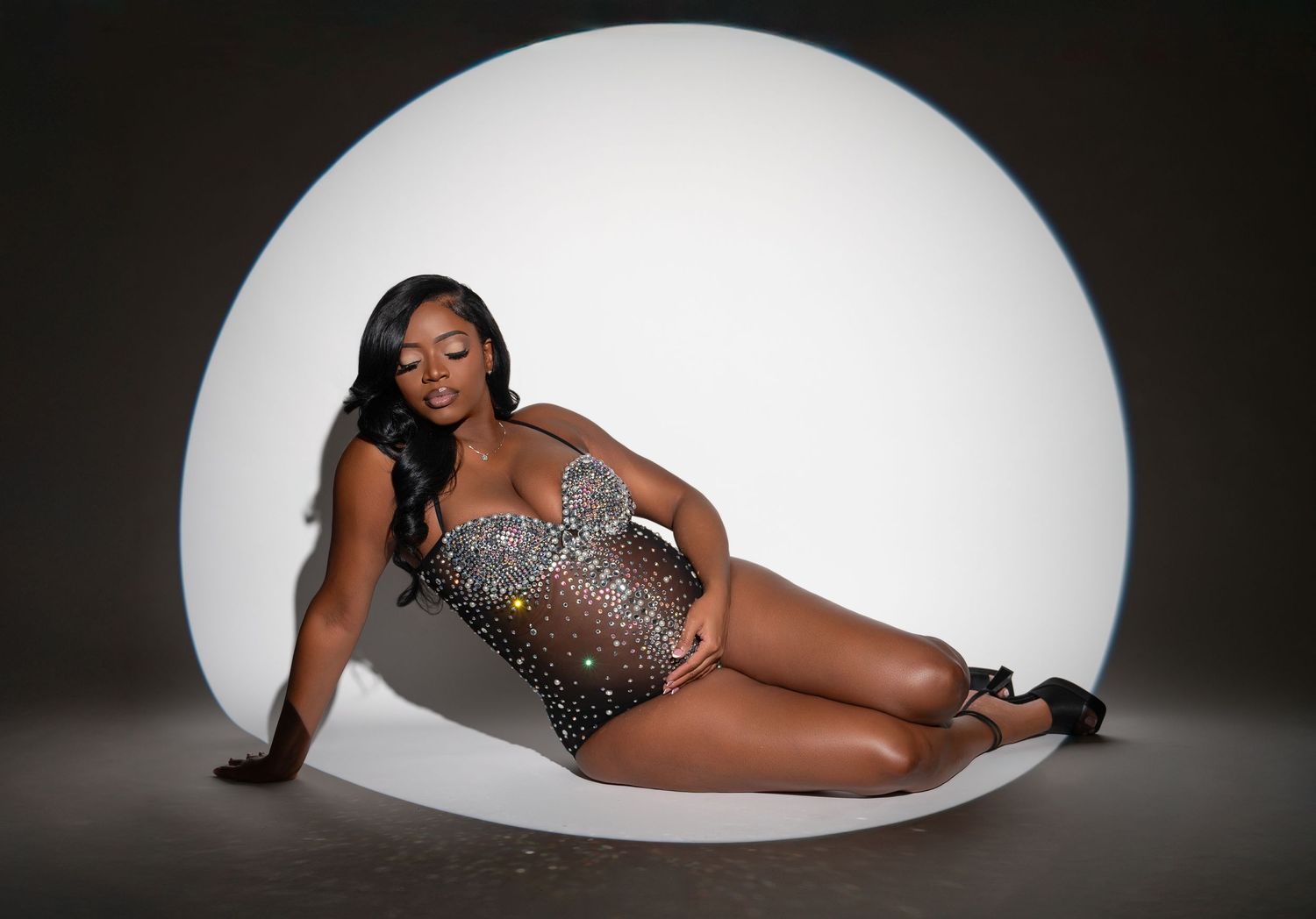 An elegant model in a sparkling bodysuit poses gracefully against a large white circular backdrop in a studio setting.