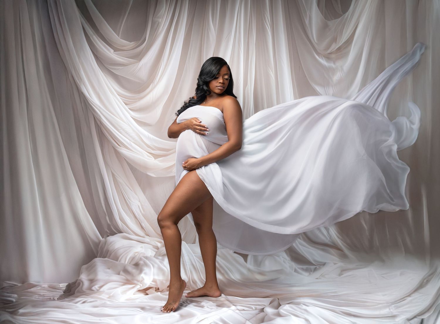 Artistic maternity photo featuring flowing white fabric against draped backdrop in elegant studio setting.