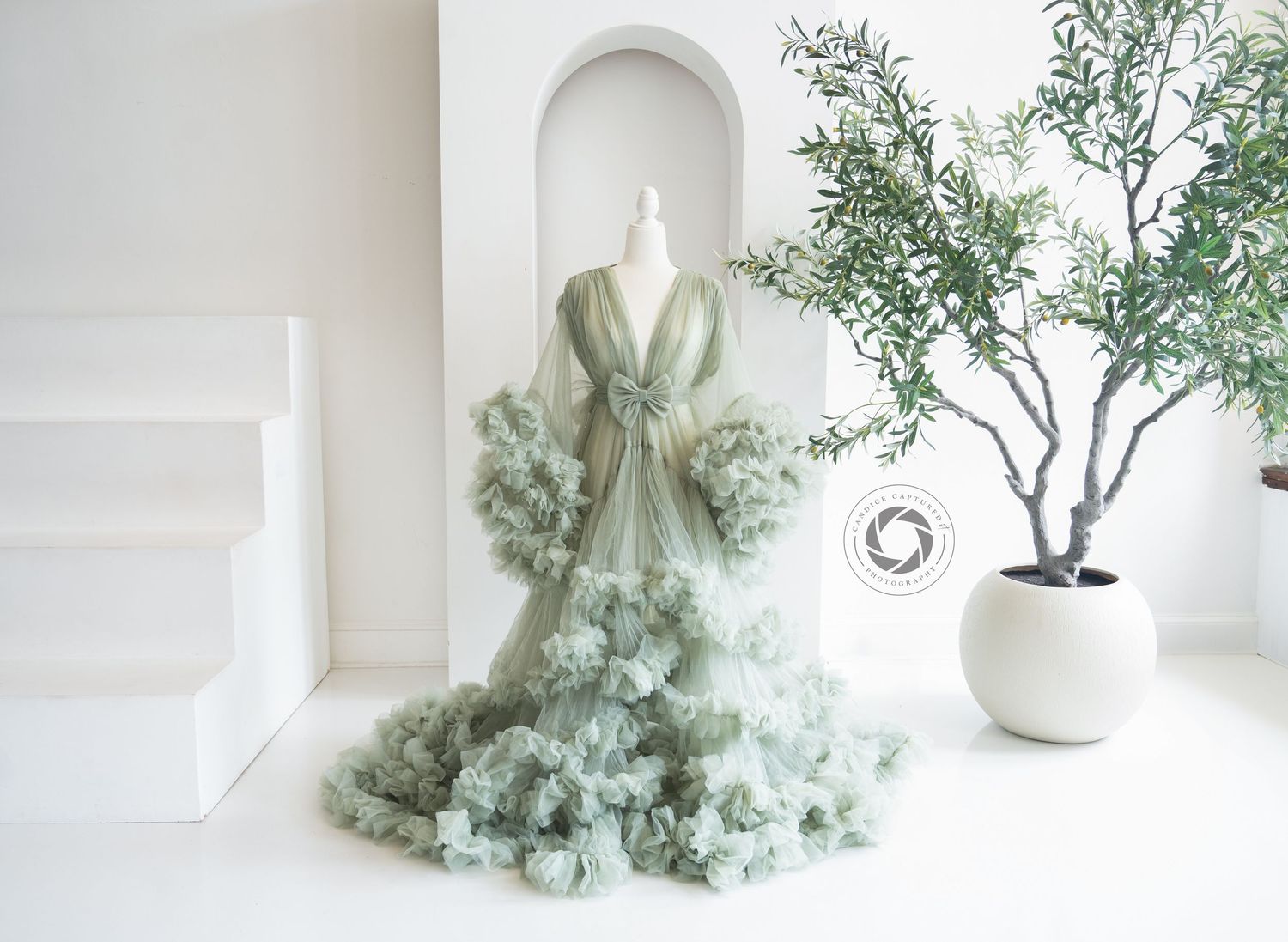 Ethereal sage green ruffled gown displayed in white minimalist room with potted olive tree decor.