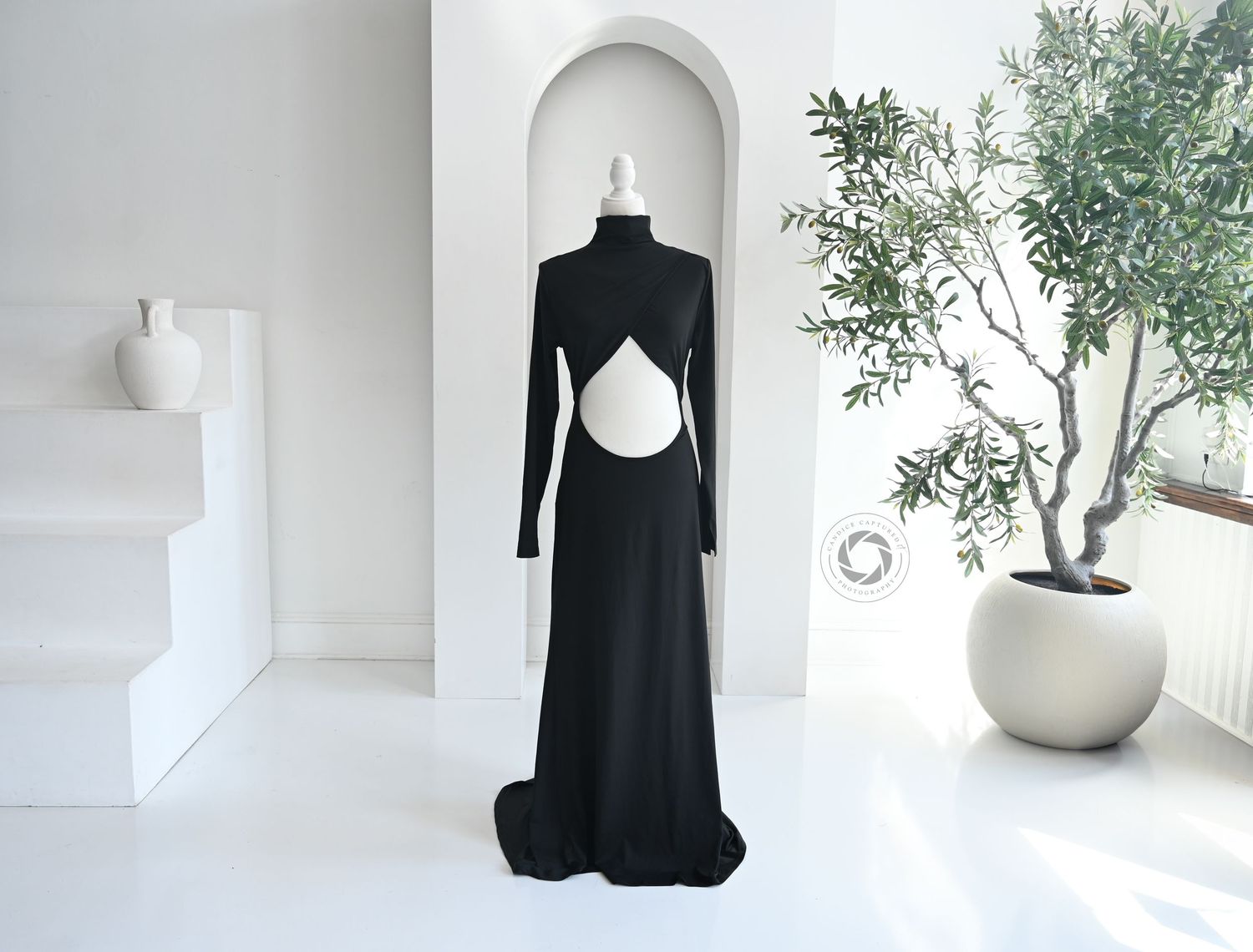 Long sleeve black evening gown with open back design displayed on mannequin in minimalist white interior setting.