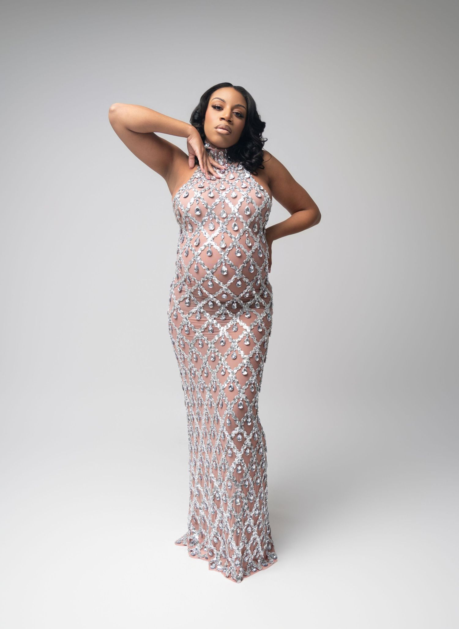 A figure poses in a form-fitting dress with geometric patterns in pink and gray tones against a white studio backdrop.