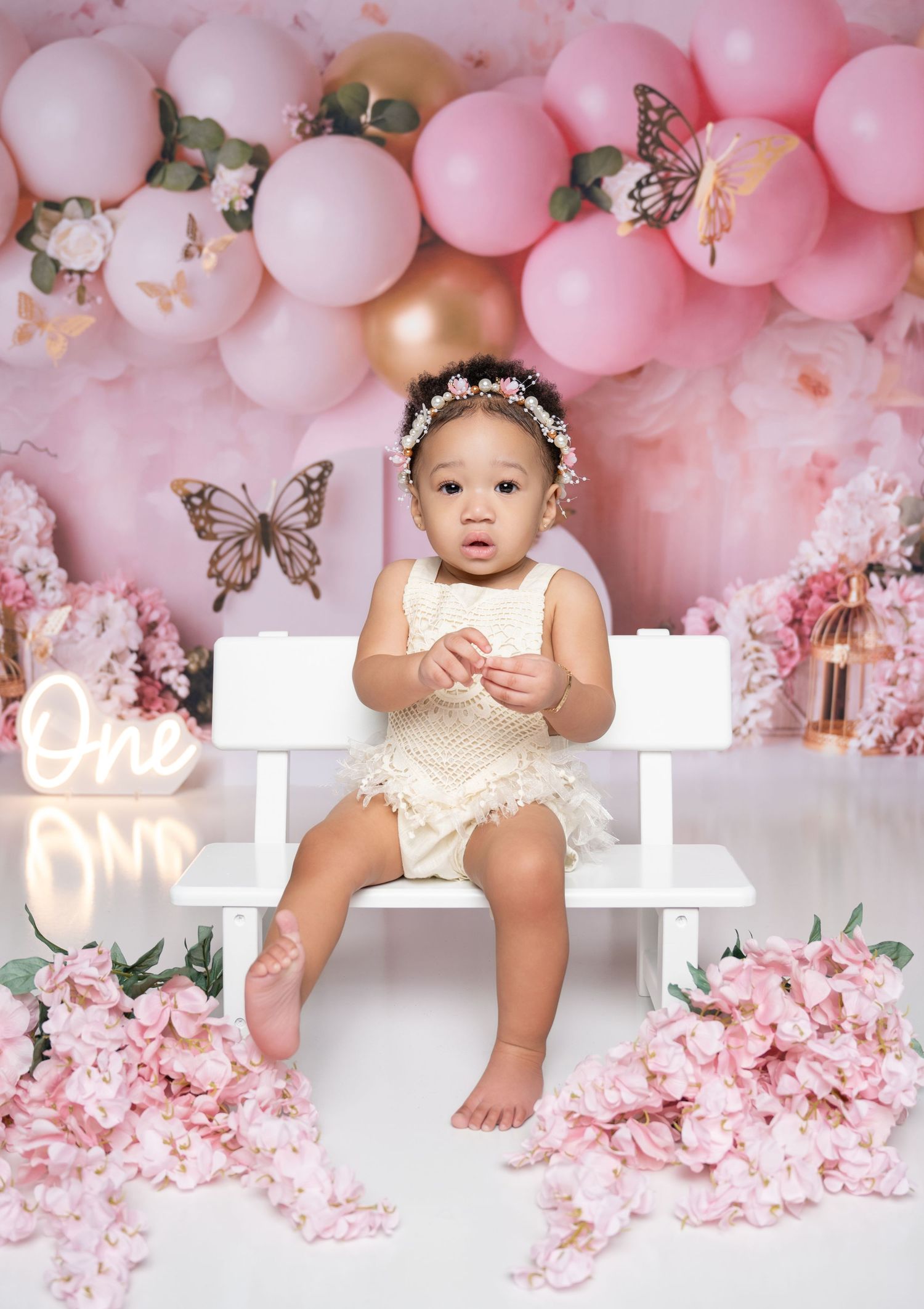 Baby girl in cream romper sits on white bench surrounded by pink balloons, butterflies, and flowers at a first birthday cake smash.