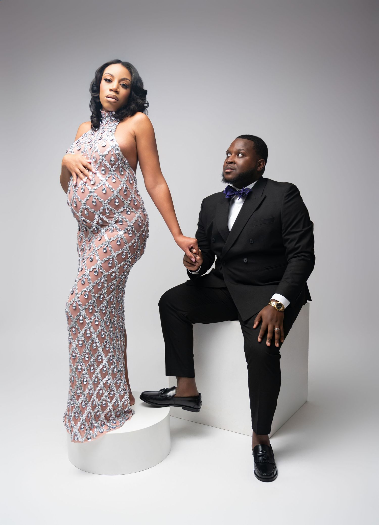 A glamorous couple poses in contrasting formal attire of a sparkly silver dress and classic black tuxedo during a studio photoshoot.