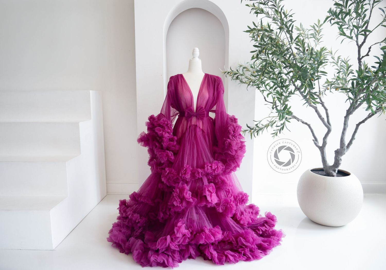 A dramatic hot pink tulle robe with voluminous ruffles displayed against a white wall next to a potted plant.