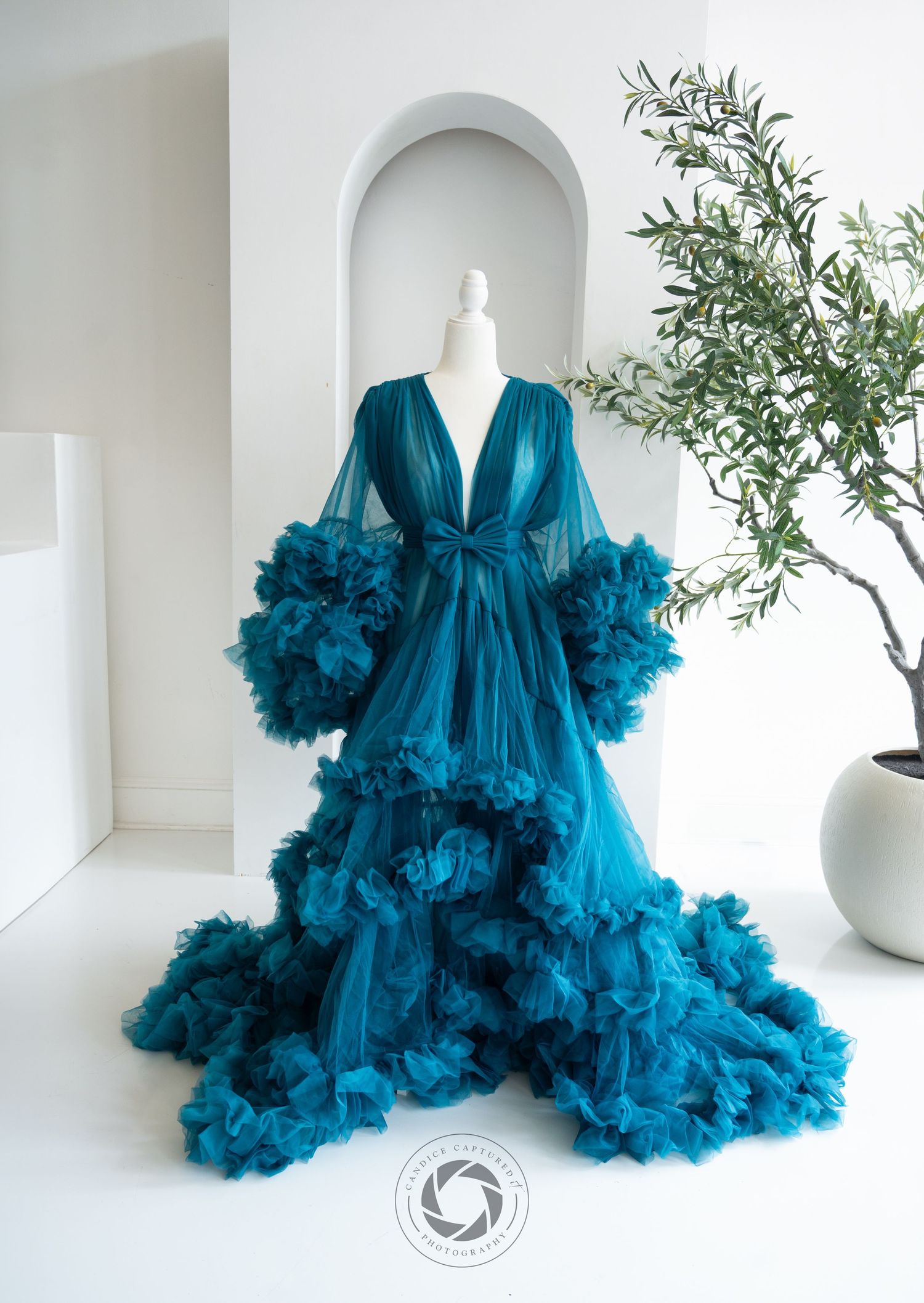 Dramatic teal ruffled tulle robe gown displayed on mannequin against white wall with arched niche and potted plant.