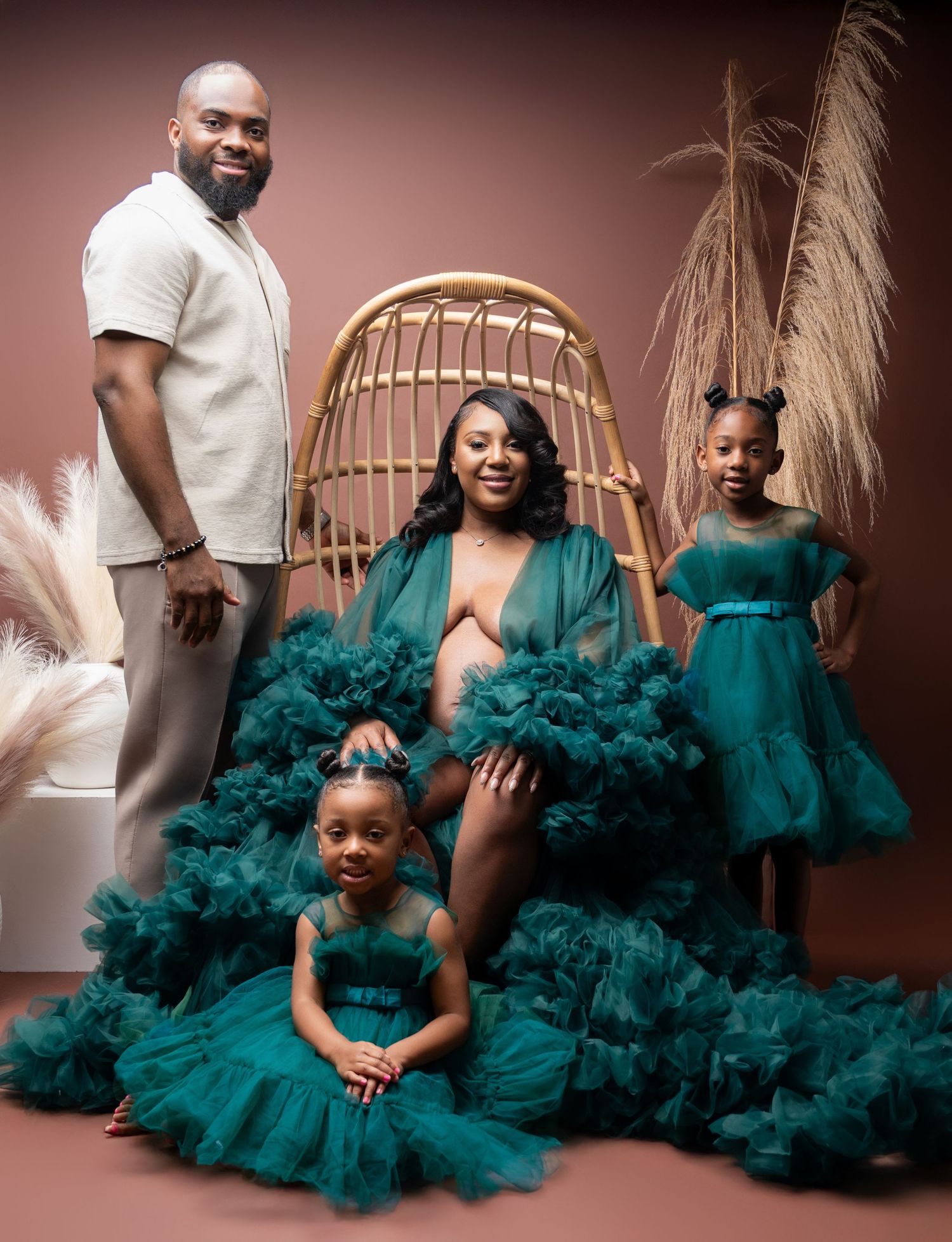 Family portrait in coordinated emerald green tulle gowns and white outfit against brown backdrop with rattan peacock chair.