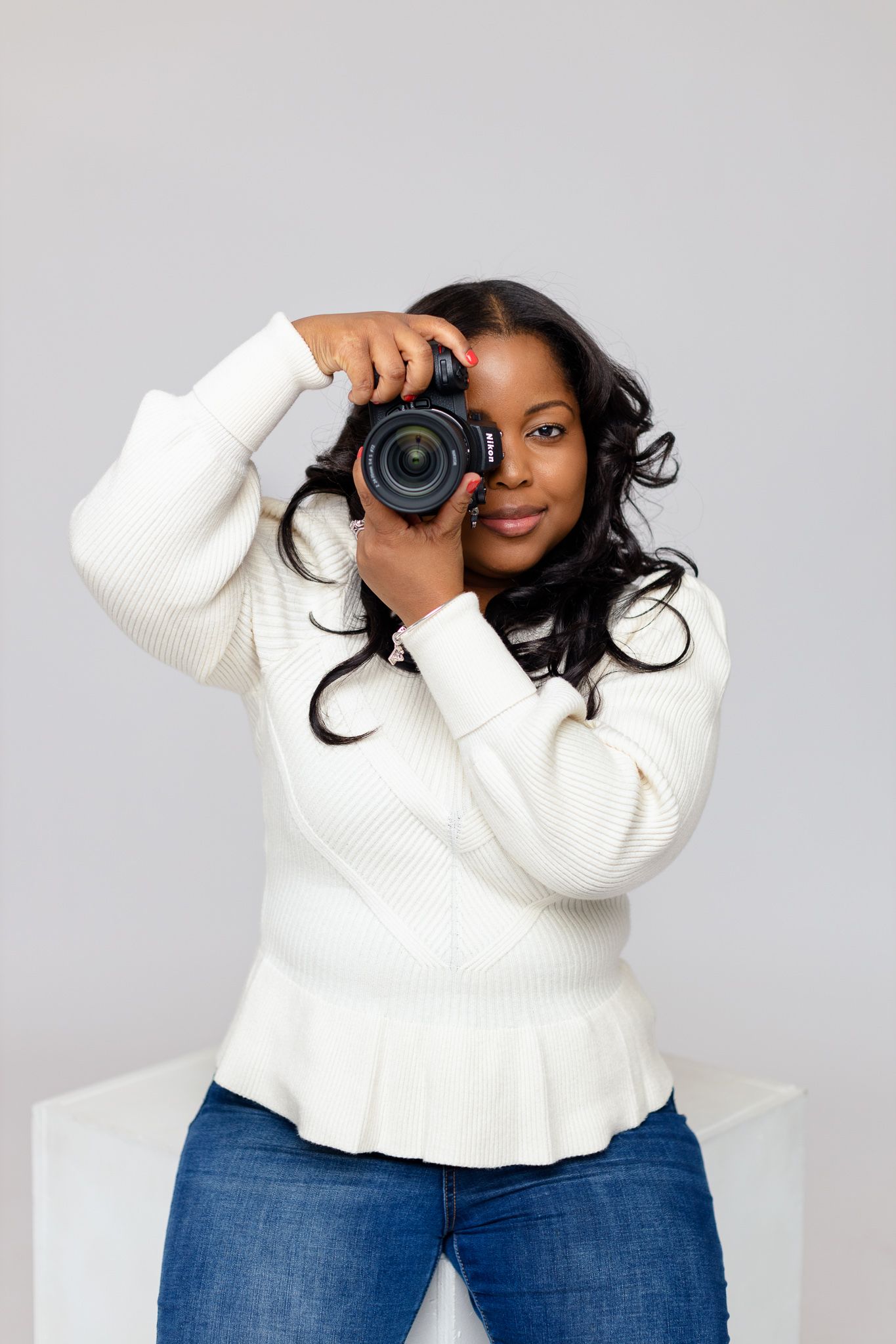 Professional photographer holding a DSLR camera while wearing a white sweater and jeans against gray background.