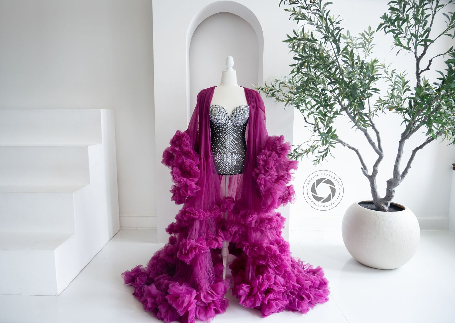 A luxurious magenta ruffled robe displayed with a silver corset on a mannequin against a white wall with potted plant.