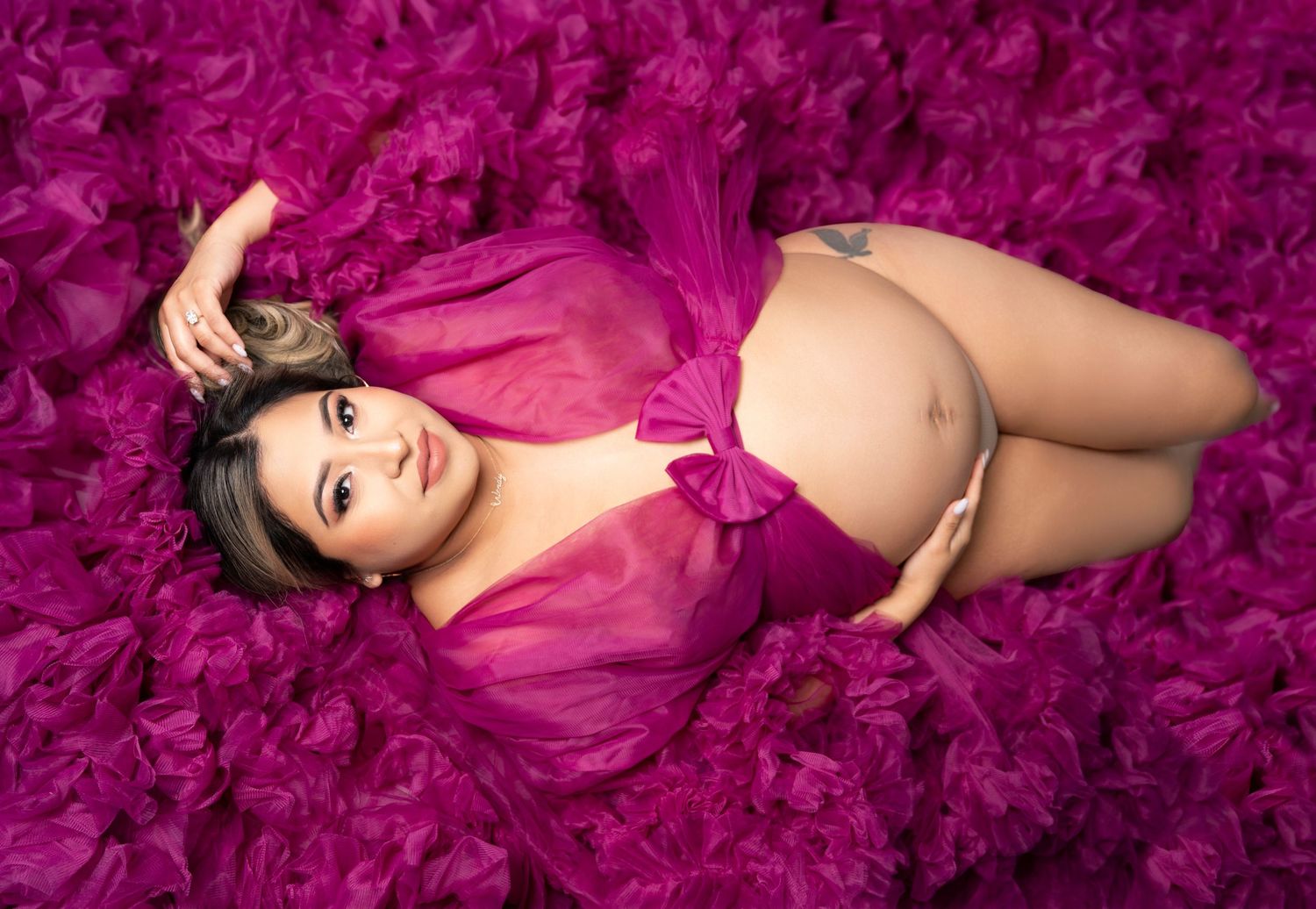 Maternity photo featuring a pregnant woman in pink sheer dress lying on vibrant fuchsia feathers.